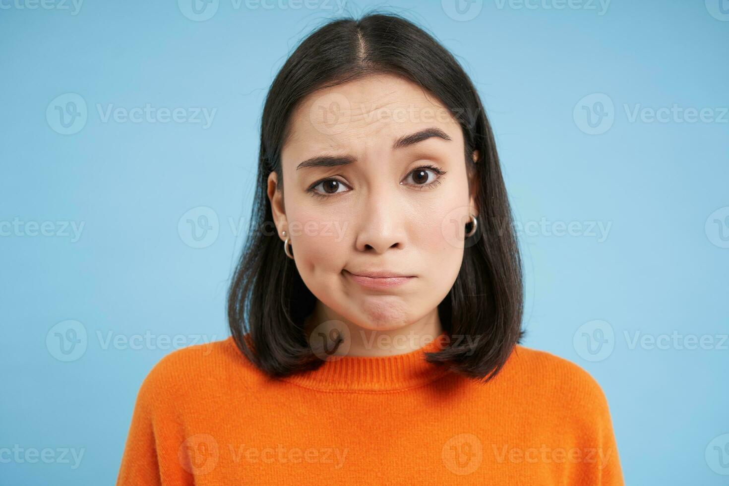 Close up of skeptical japanese girl, shrugs shoulders and looks confused, puzzled, stands over blue studio background photo