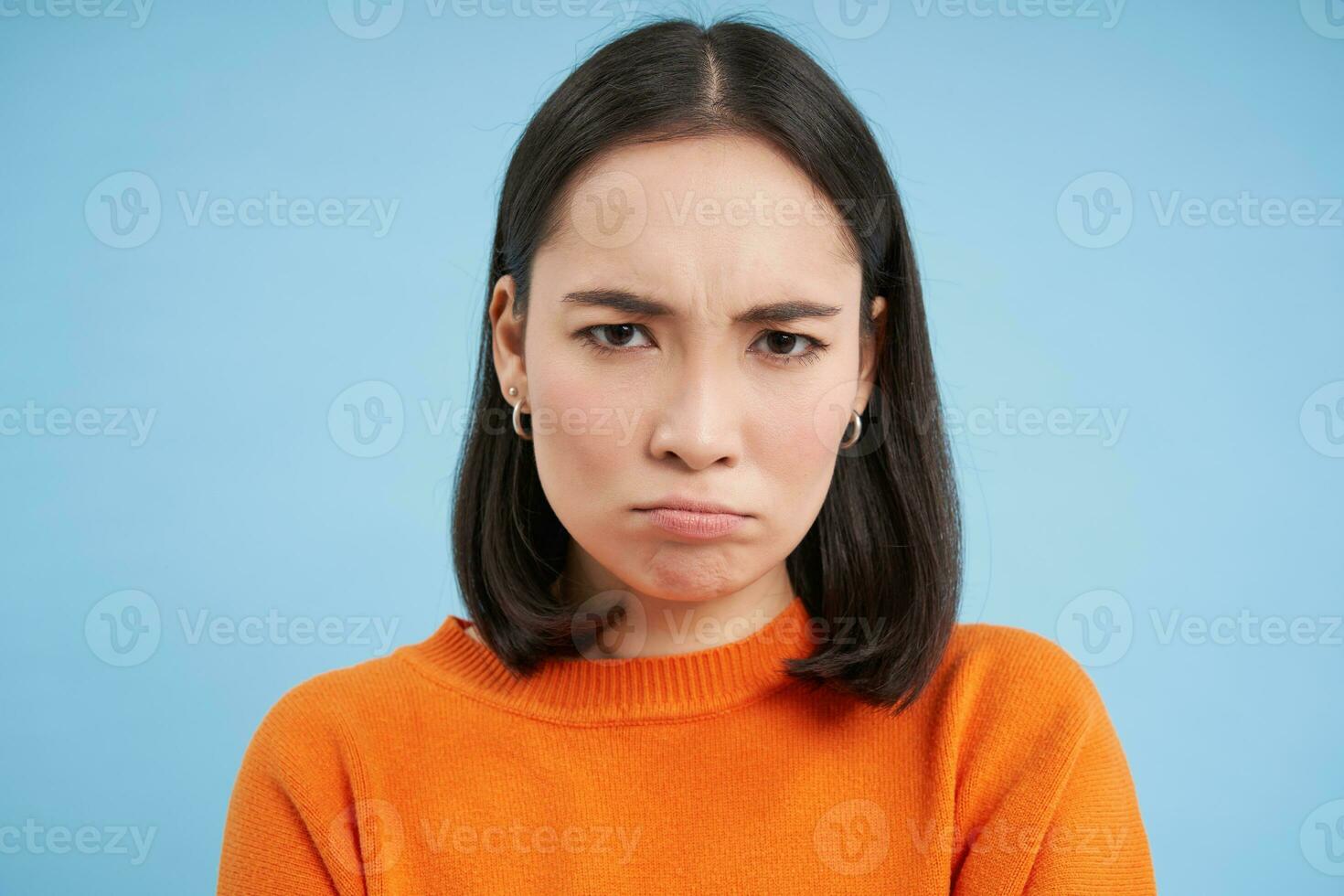 Close up of sad and gloomy japanese girl, sulking and looking upset, stands over blue background photo