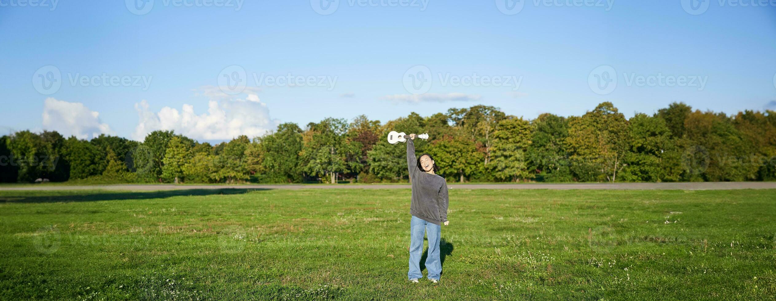 Excited young woman lift her ukulele up over head and smiles, posing with musical instrument in green field park photo