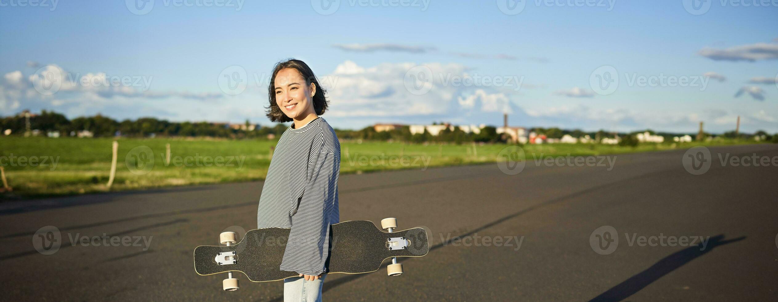 Vertical shot of carefree asian girl with longboard. Young woman skater holding cruiser on her shoulders and walking on road, skateboarding photo