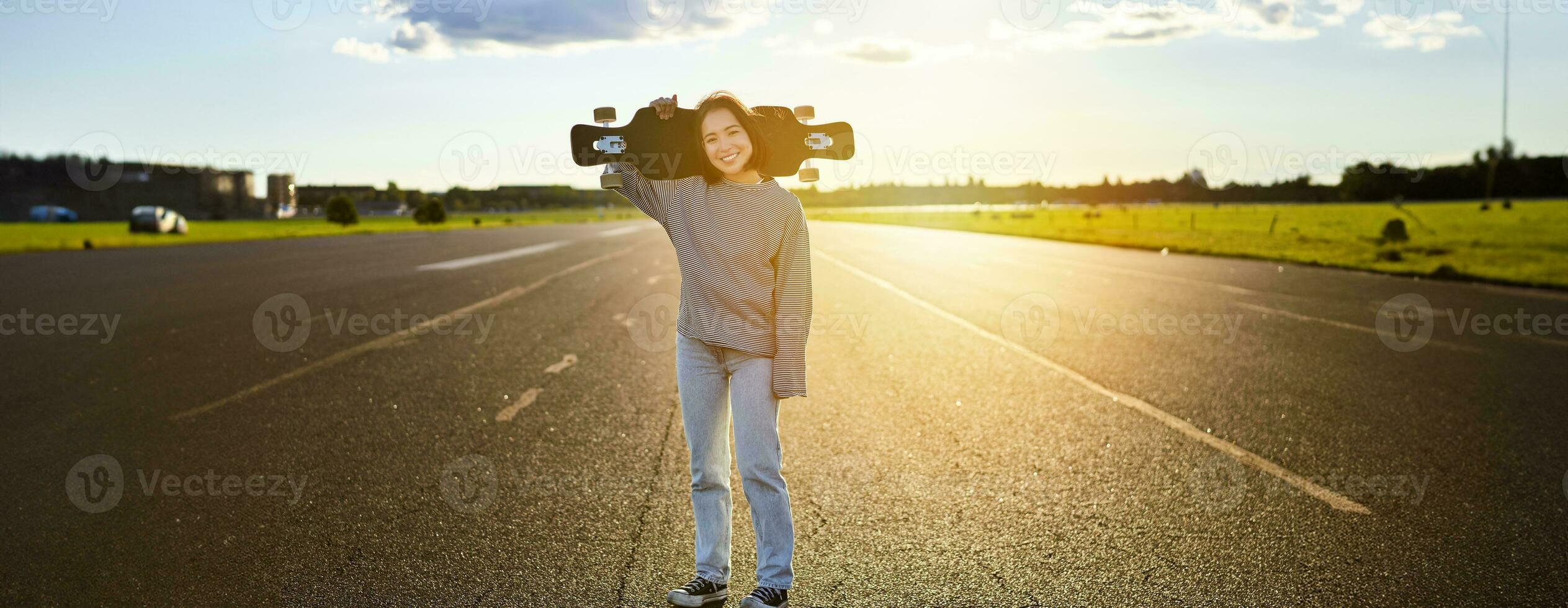 Asian girl with skateboard standing on road during sunset. Skater posing with her long board, cruiser deck during training photo