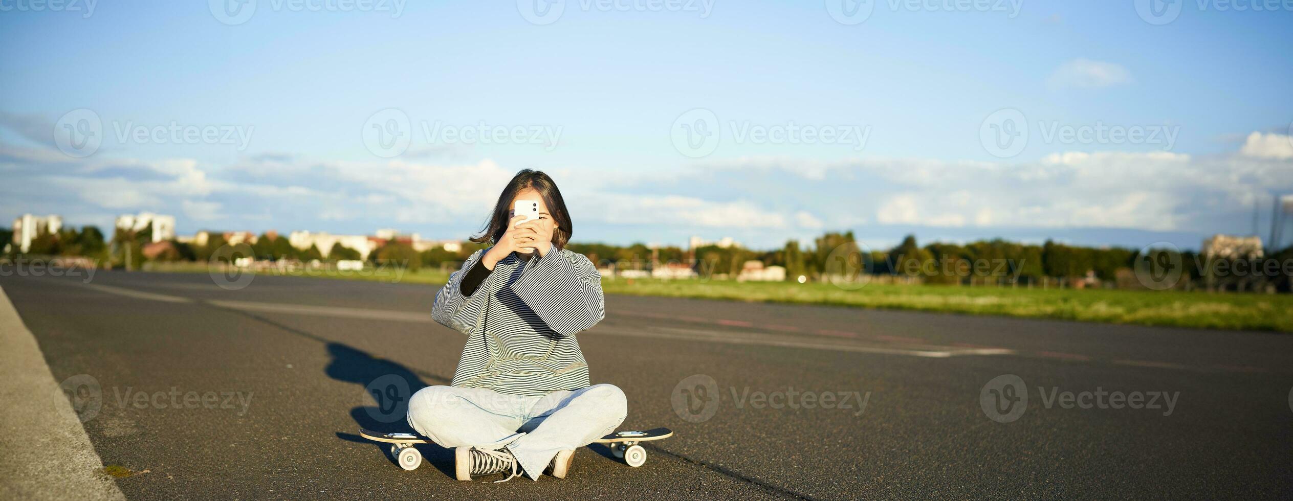 Hipster teen girl sitting on her skateboard, taking photos on smartphone. Asian woman skater sits on longboard and photographing on mobile phone
