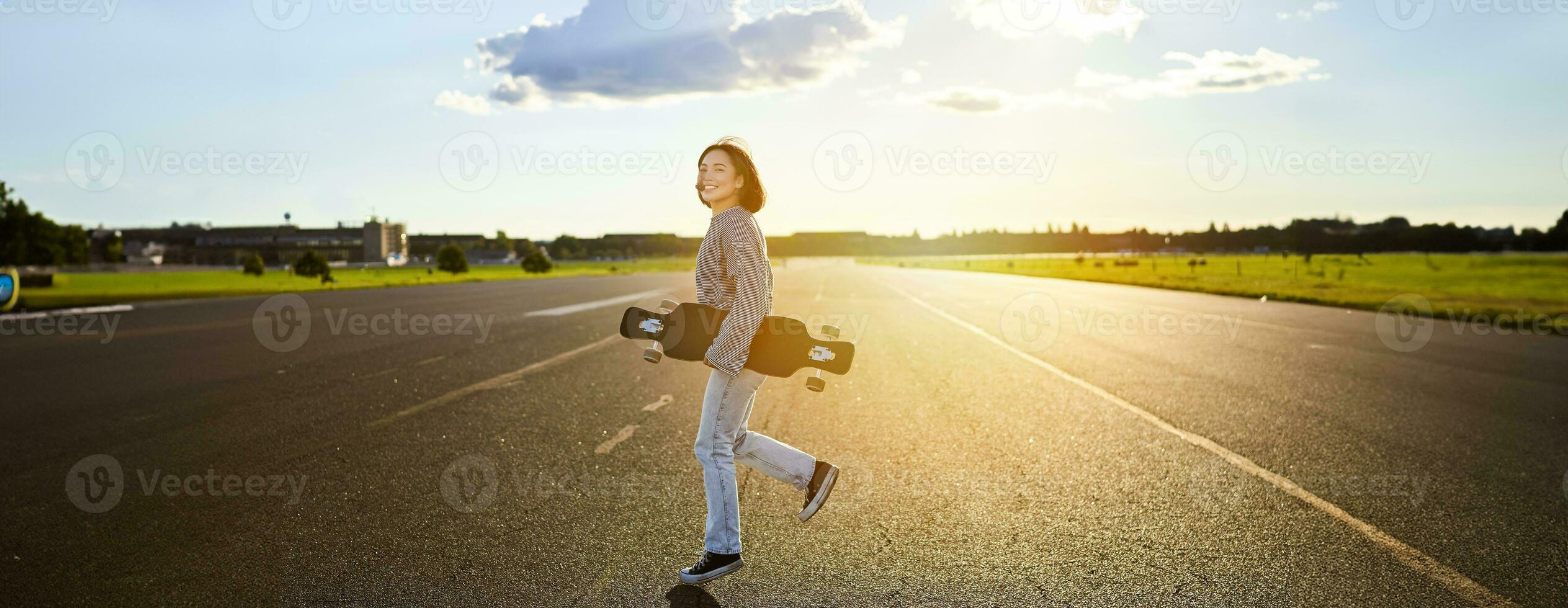 asiático niña con patineta en pie en la carretera durante puesta de sol. patinador posando con su largo junta, crucero cubierta durante formación foto