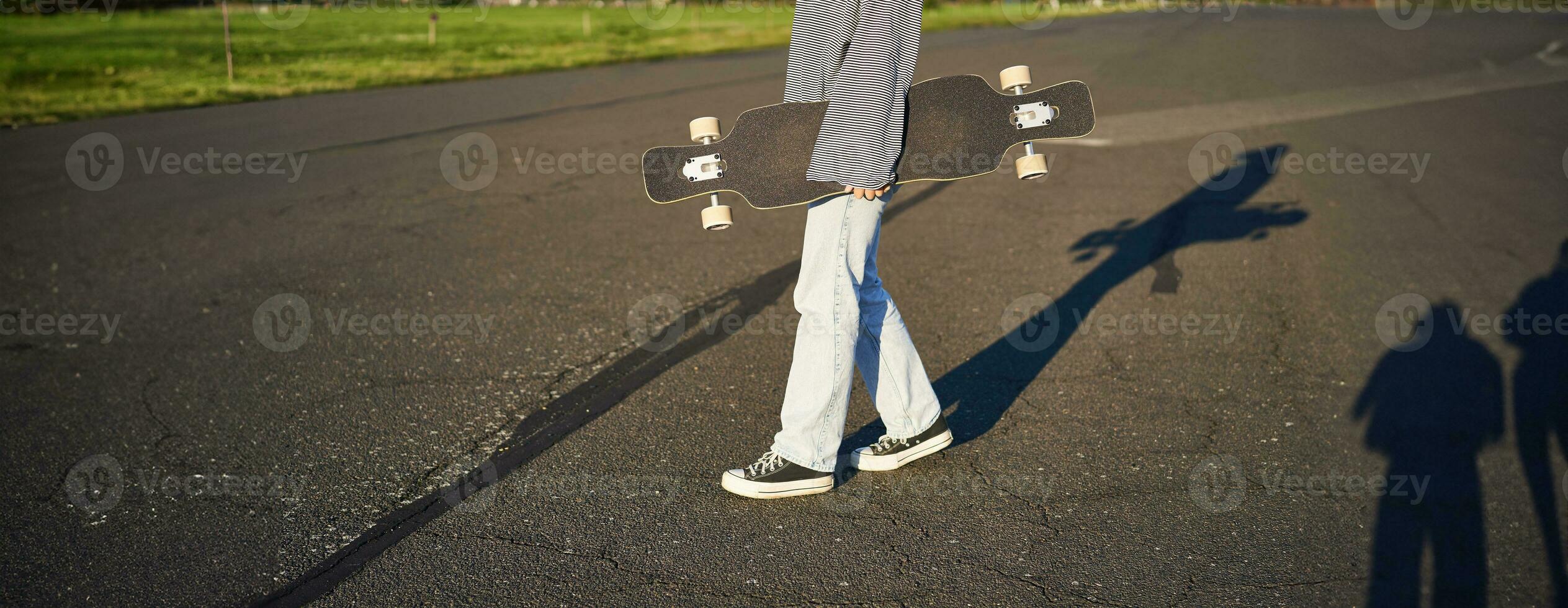 Cropped shot of teen girl body, holding cruiser longboard in hand, walking in sneakers on road in jeans and sweatshirt. Young woman skater with skateboard photo