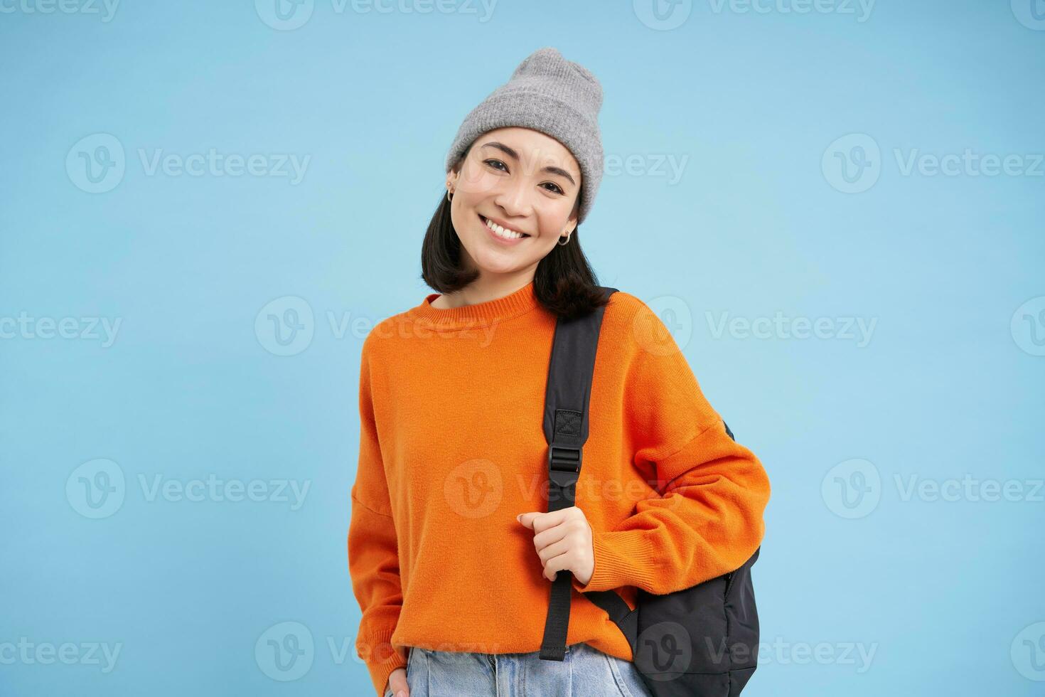Stylish young asian woman in warm hat, walking with backpack, going somewhere with bag, standing over blue studio background photo