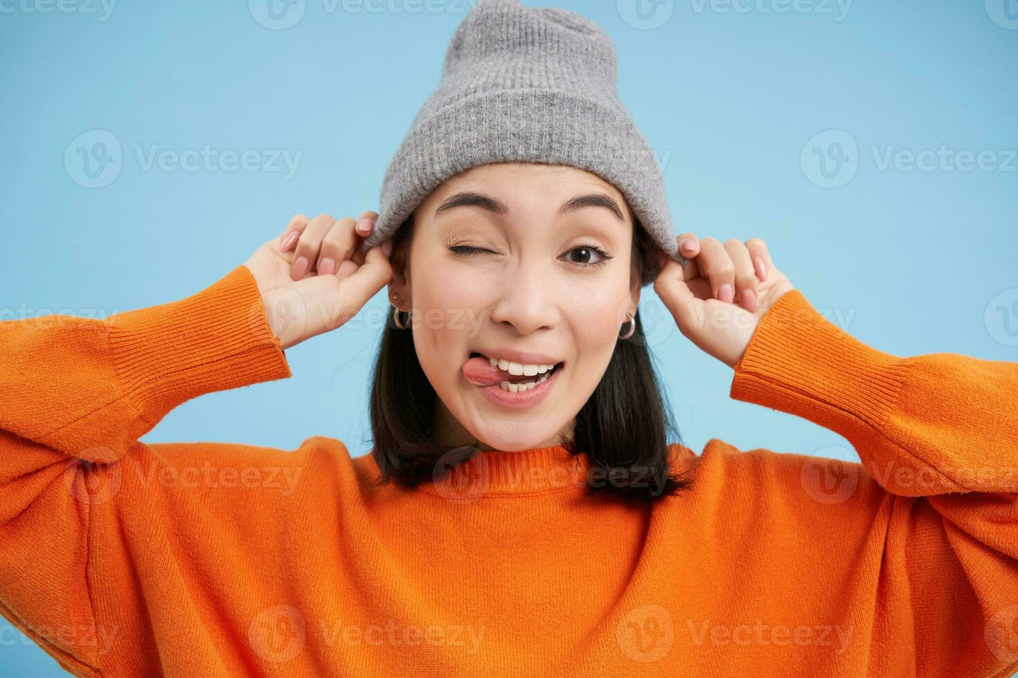 Close up portrait of Asian female model puts on hat and winks at you, looking happy, going outside, standing over blue background photo