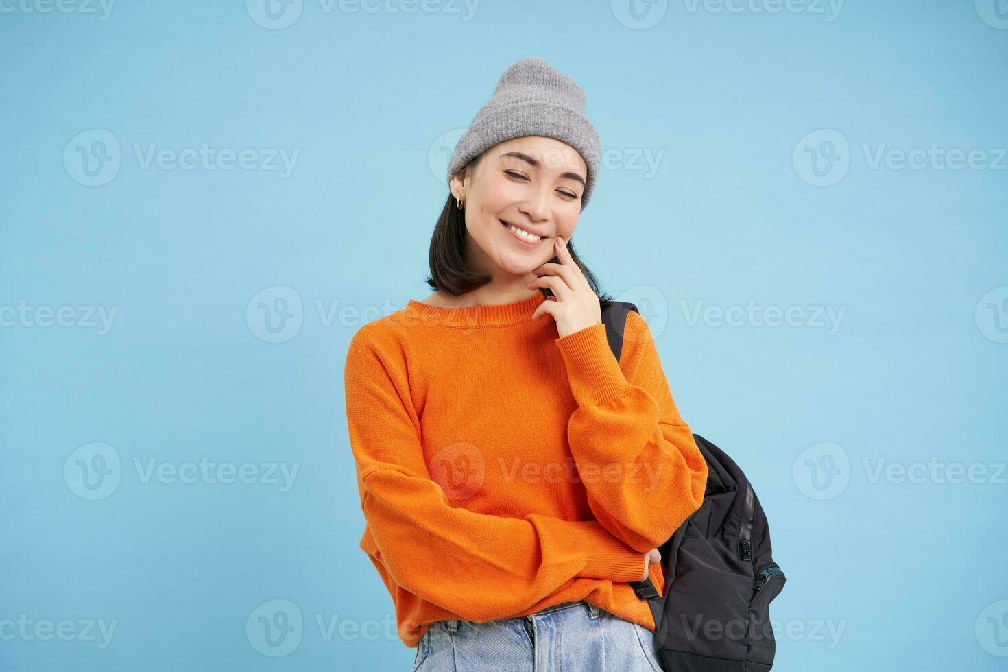 Modern college girl in hat, holds backpack on shoulder, smiles and laughs, stands over studio background photo