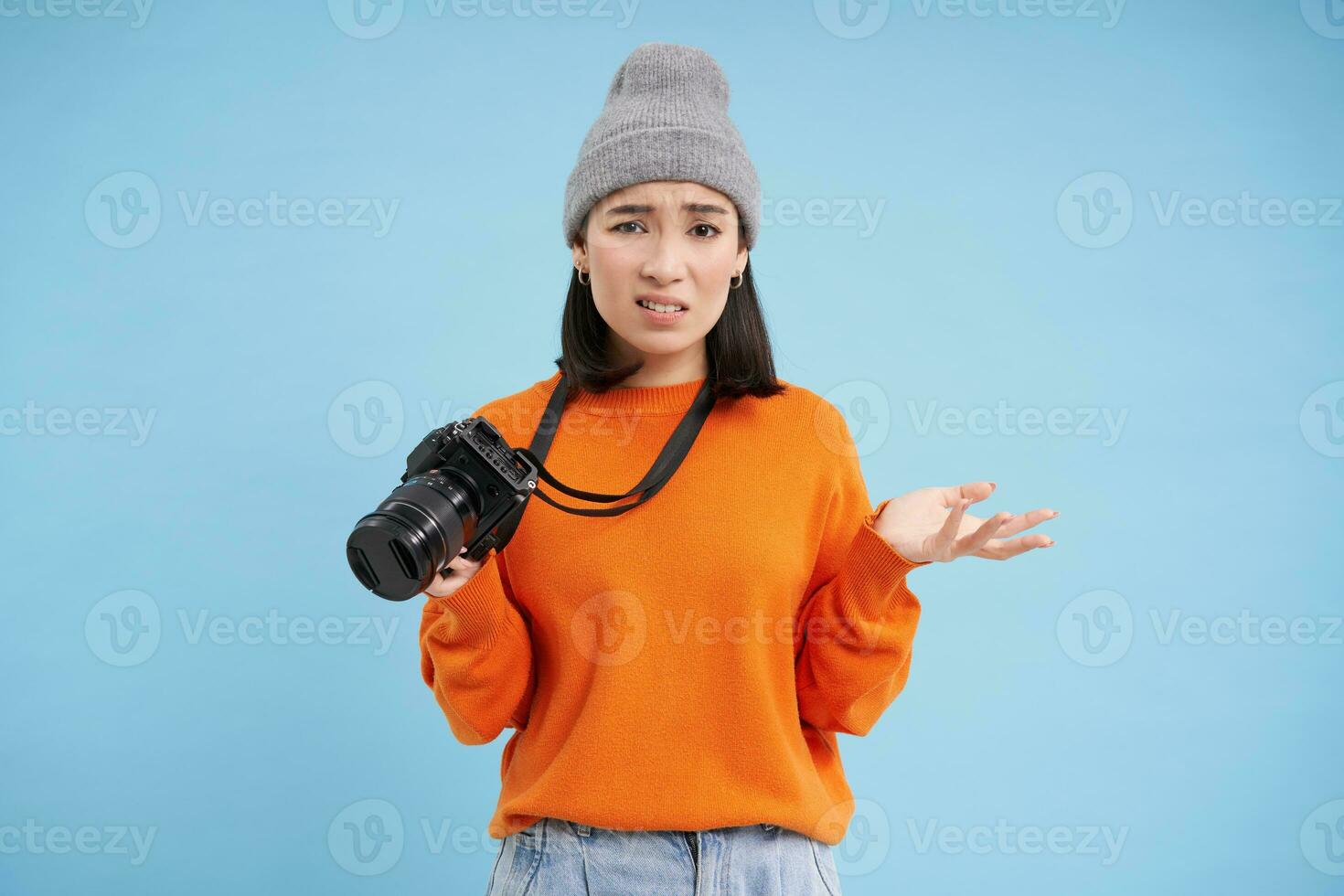 Young confused asian girl in hat, holds digital camera and shrugging shoulders, clueless how to take pictures on digicam, blue background photo