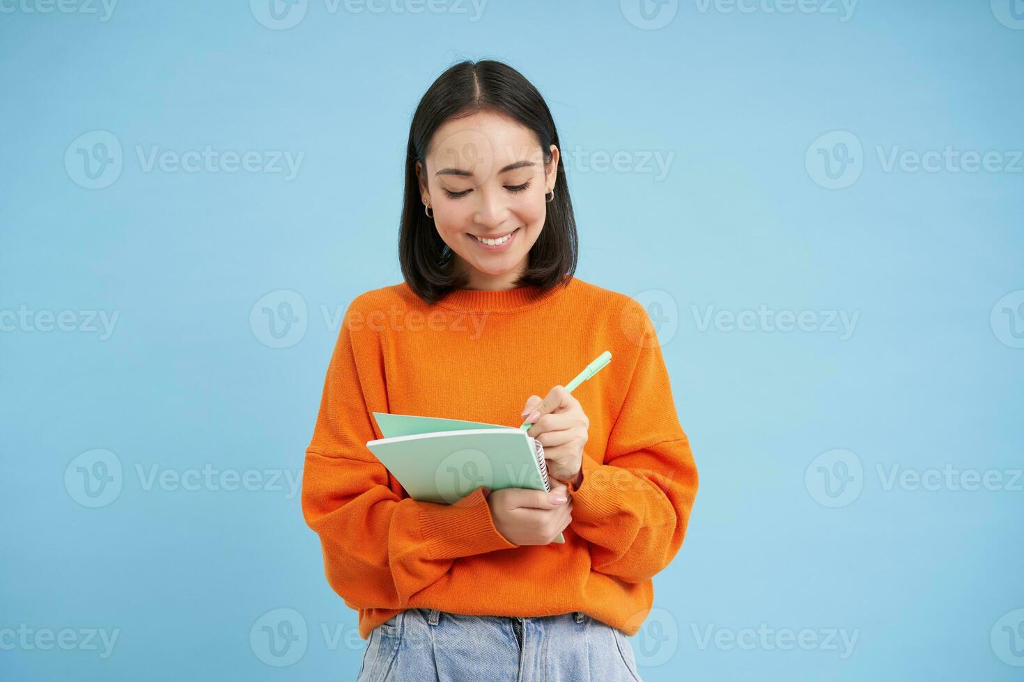Smiling cute girl student, making notes, writing down homework, studying, standing over blue studio background photo