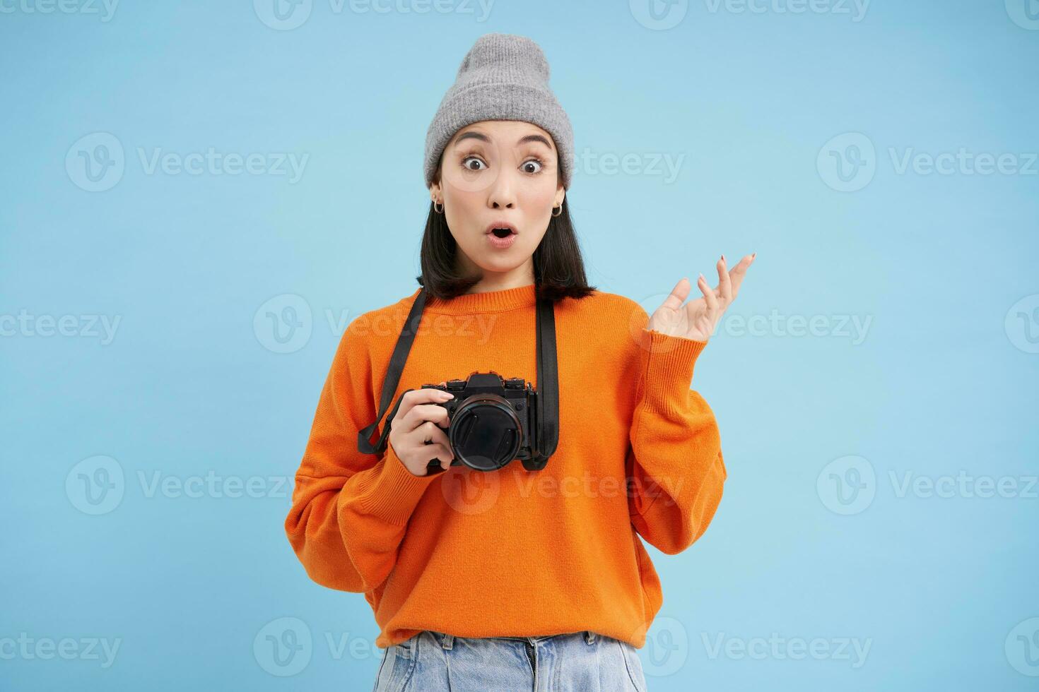 Surprised asian girl in hat, holds digital camera, looks amazed and impressed, poses over blue studio background with space for banner photo