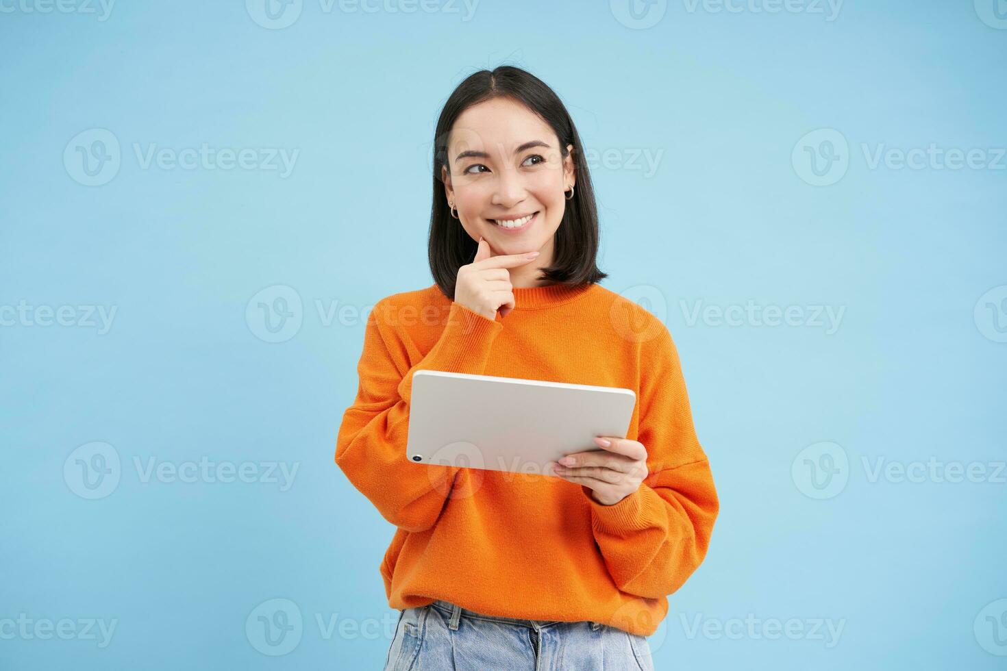 Woman with digital tablet thinking, looking aside with thoughtful face expression, standing over blue background photo