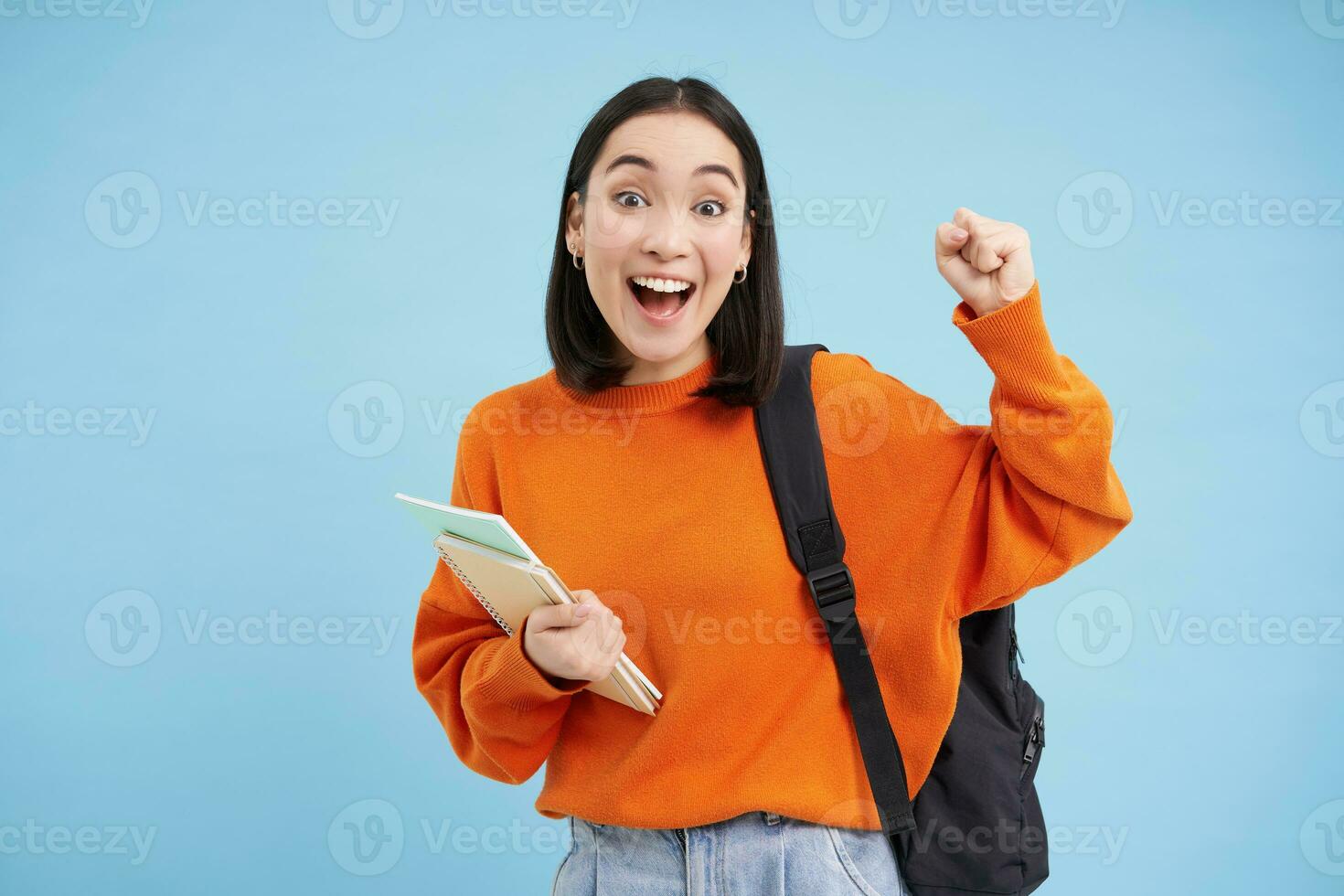 Enthusiastic girl student, celebrating, wearing backpack and holding notebooks, college homework, standing over blue background photo