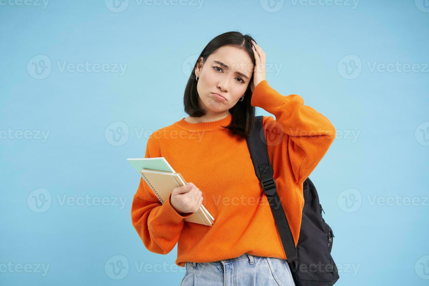 triste niña alumno, toques su cabeza y se enfurruña, mira cansado después colega, sostiene mochila y cuadernos, azul antecedentes foto