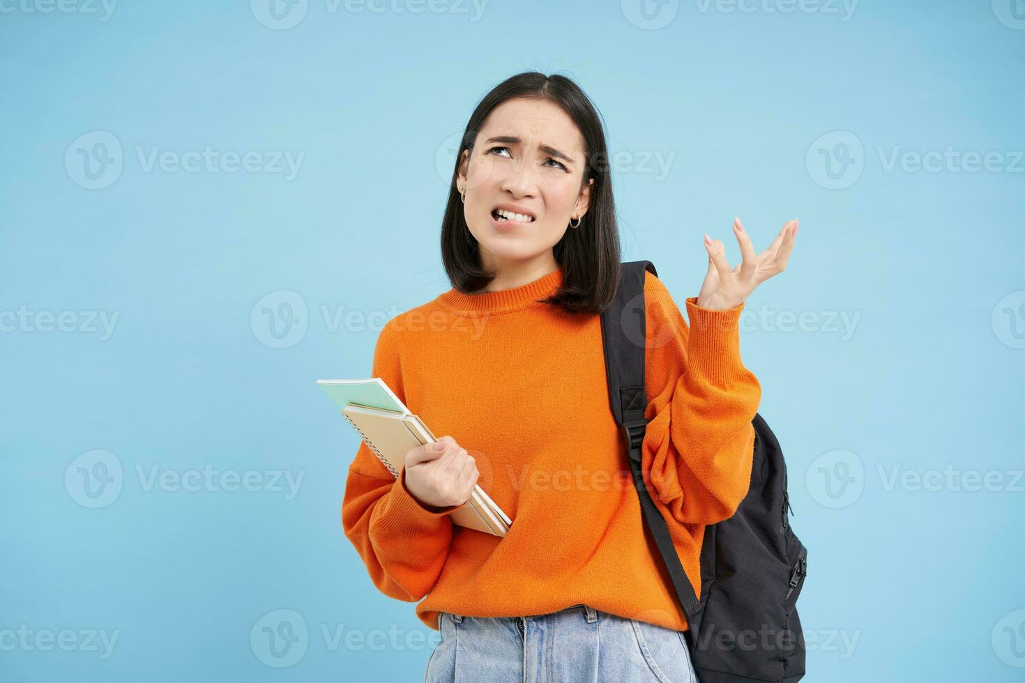 irritado joven asiático mujer, estudiante complan, batidos mano y mira decepcionado, soportes con mochila y cuadernos, azul antecedentes foto