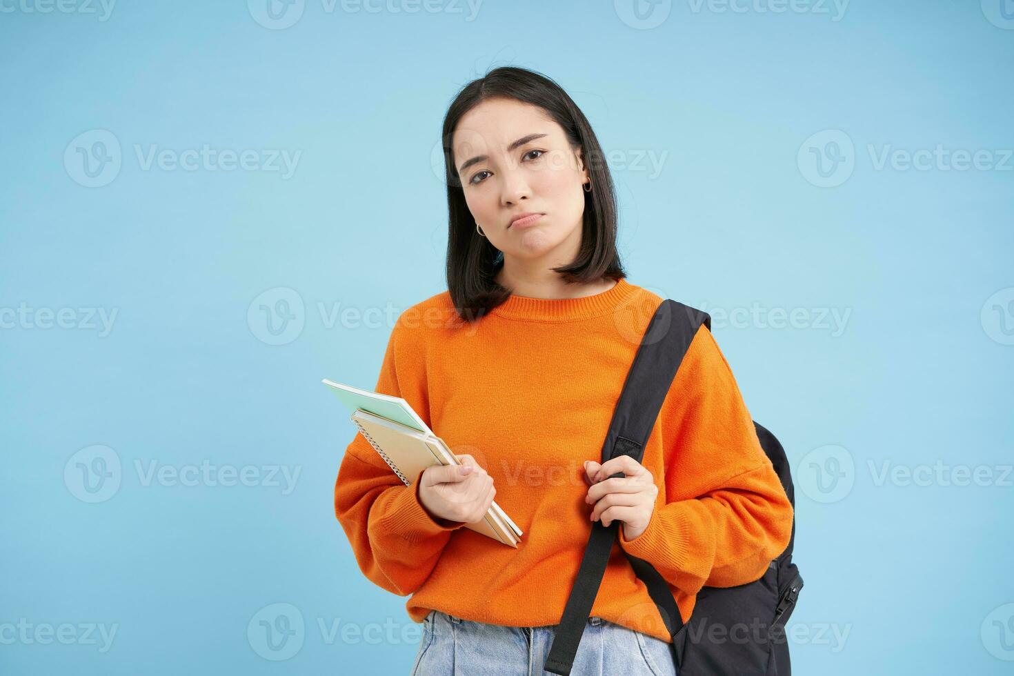 triste malhumorado asiático alumno, niña con mochila y cuadernos, mirando decepcionado, en pie terminado azul antecedentes foto