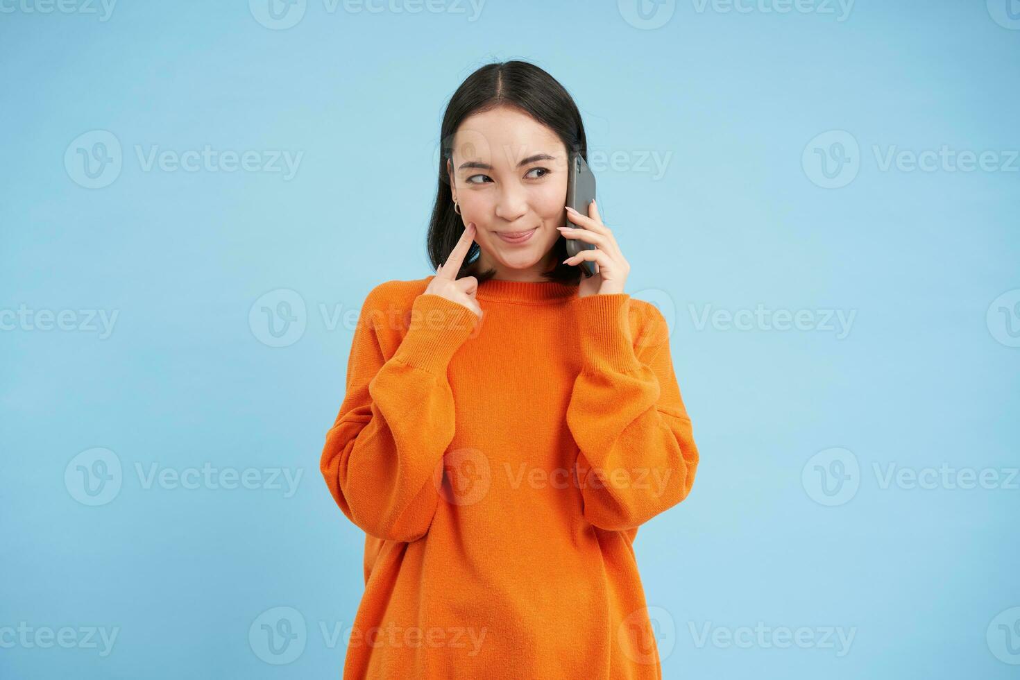 Young woman with thoughtful look, answers phone call, talks on mobile and smiling, thinking with pleased face, standing over blue background photo