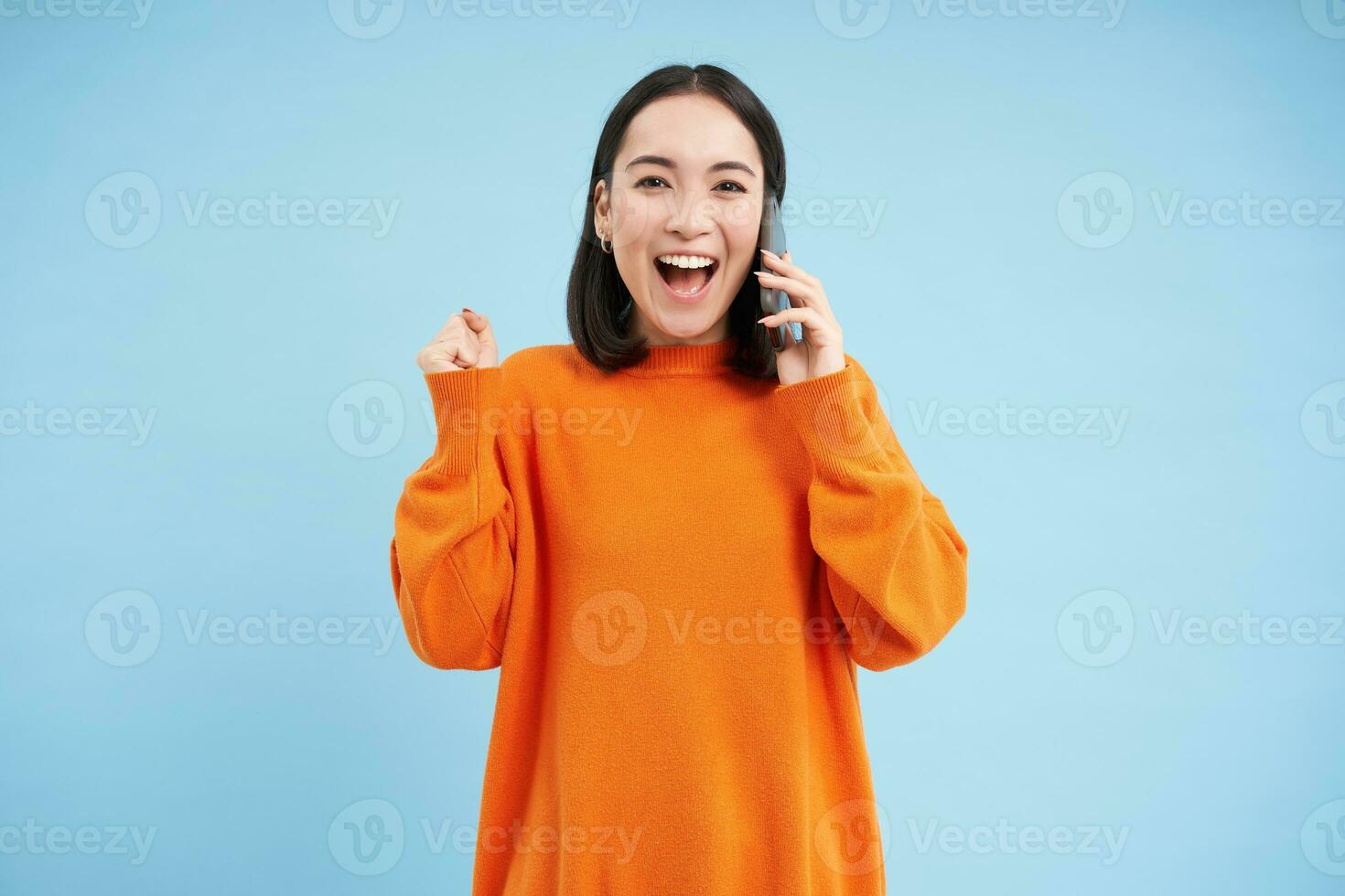 Happy asian girl receives excellent news over the phone, holds mobile and does fist pump, celebrates victory, achievement, blue studio background photo
