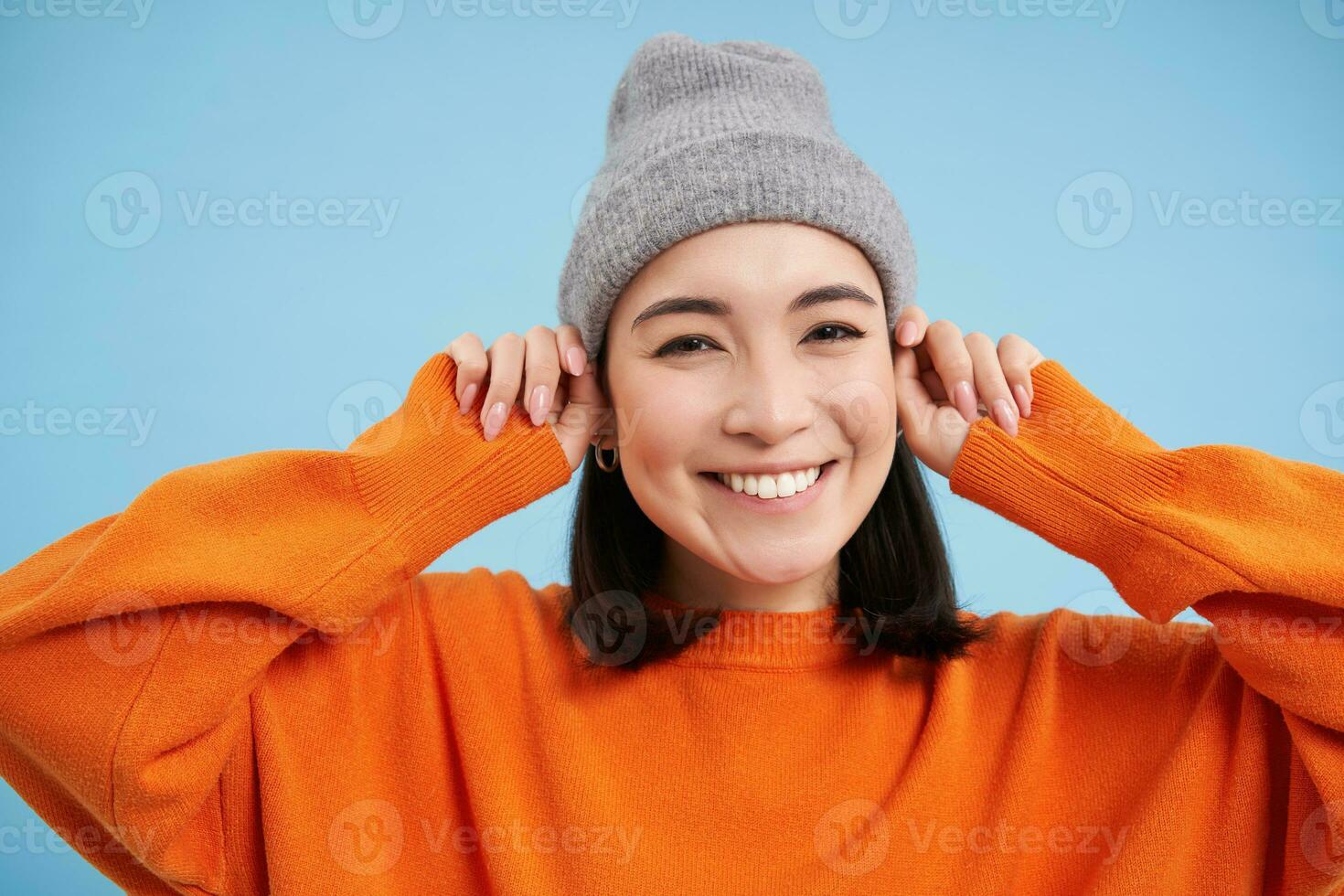 Close up portrait of happy, stylish korean girl puts on warm hat, smiles and looks joyful, stands against blue studio background photo