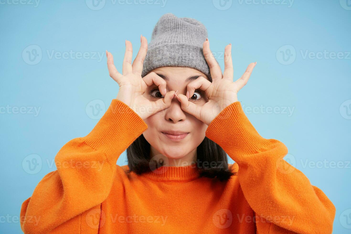 Close up portrait of funny Chinese girl, looks through hand glasses with surprised face expression, standing over blue background photo