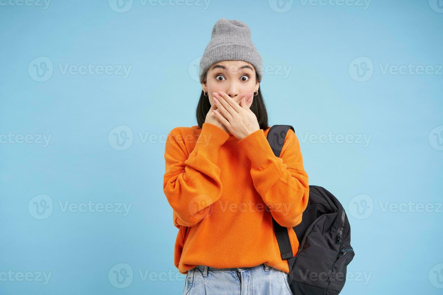 Portrait of korean girl in hat, covers her mouth with hands, looks shocked, amazed by gossips, stands over blue background photo