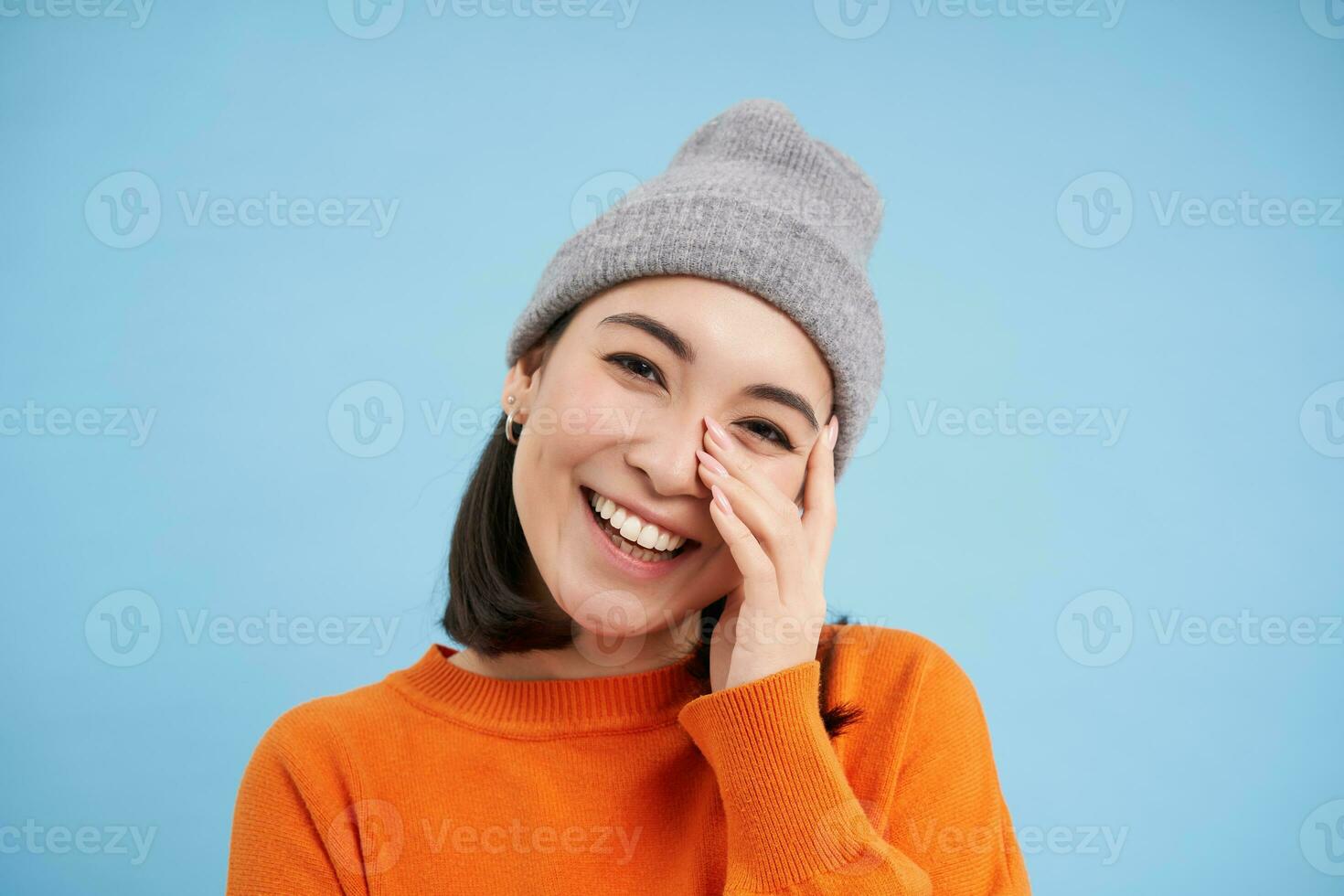 Close up portrait of smiling asian woman in warm hat, looking happy and cute at camera, has clear natural skin, stands over blue background photo