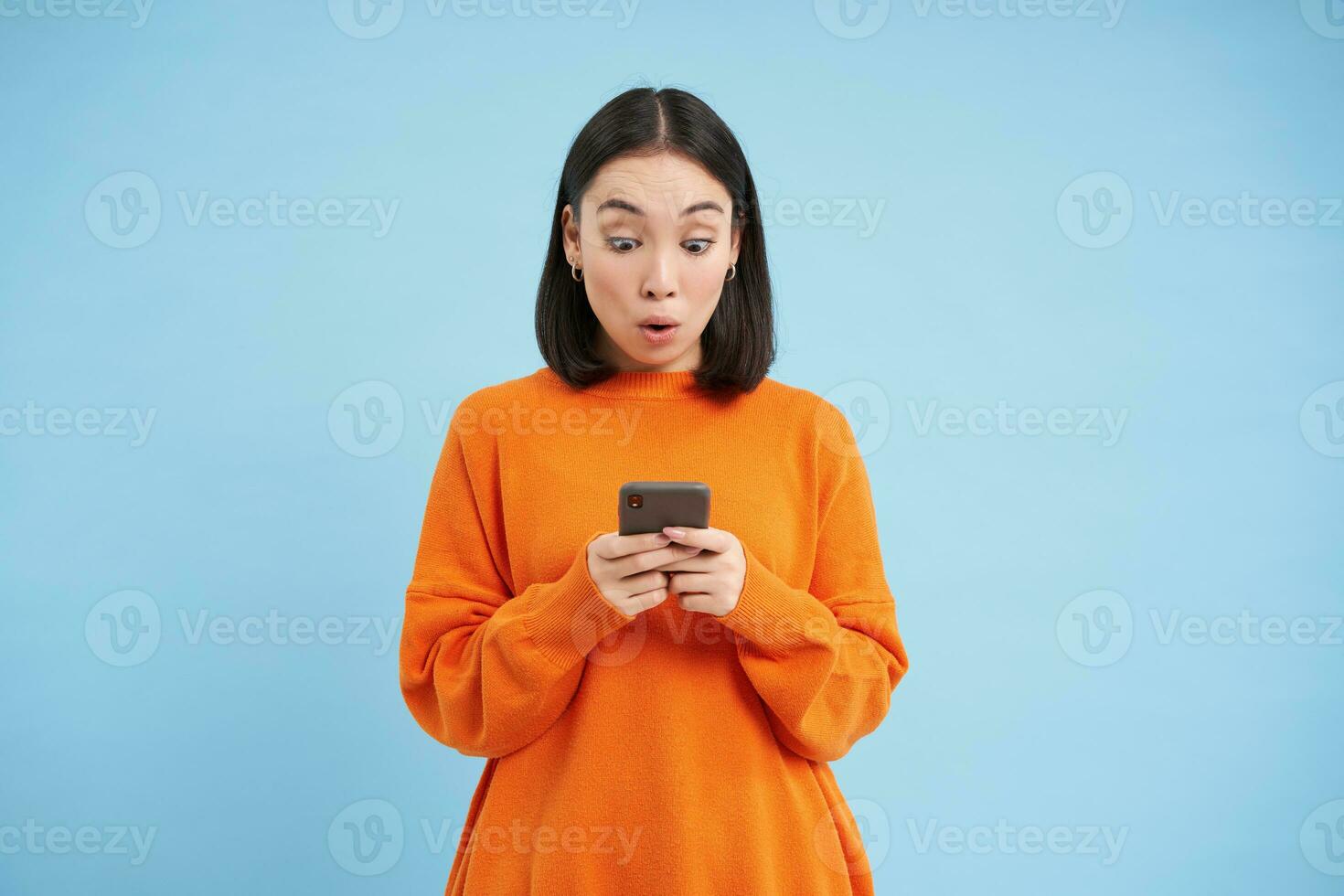 joven mujer mira asombrado a su Teléfono móvil, lee emocionante noticias, encontró bueno acuerdo en línea en móvil solicitud, en pie terminado azul antecedentes foto