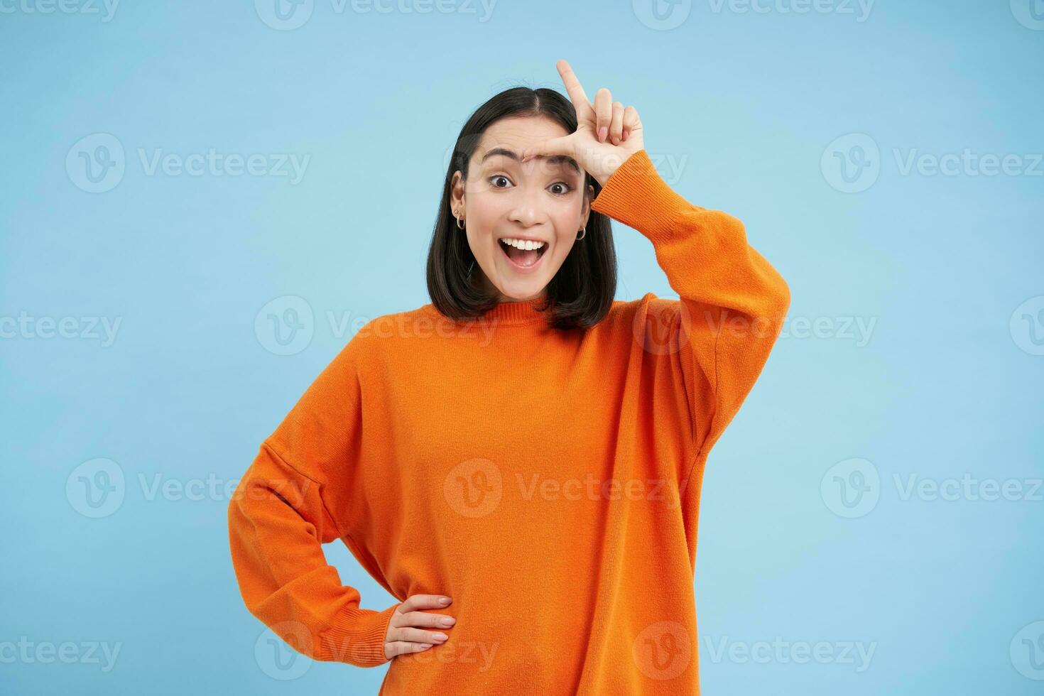Enthusiastic korean girl, shows L letter on forehead and laughs, mocks someone who lost, stands over blue background photo