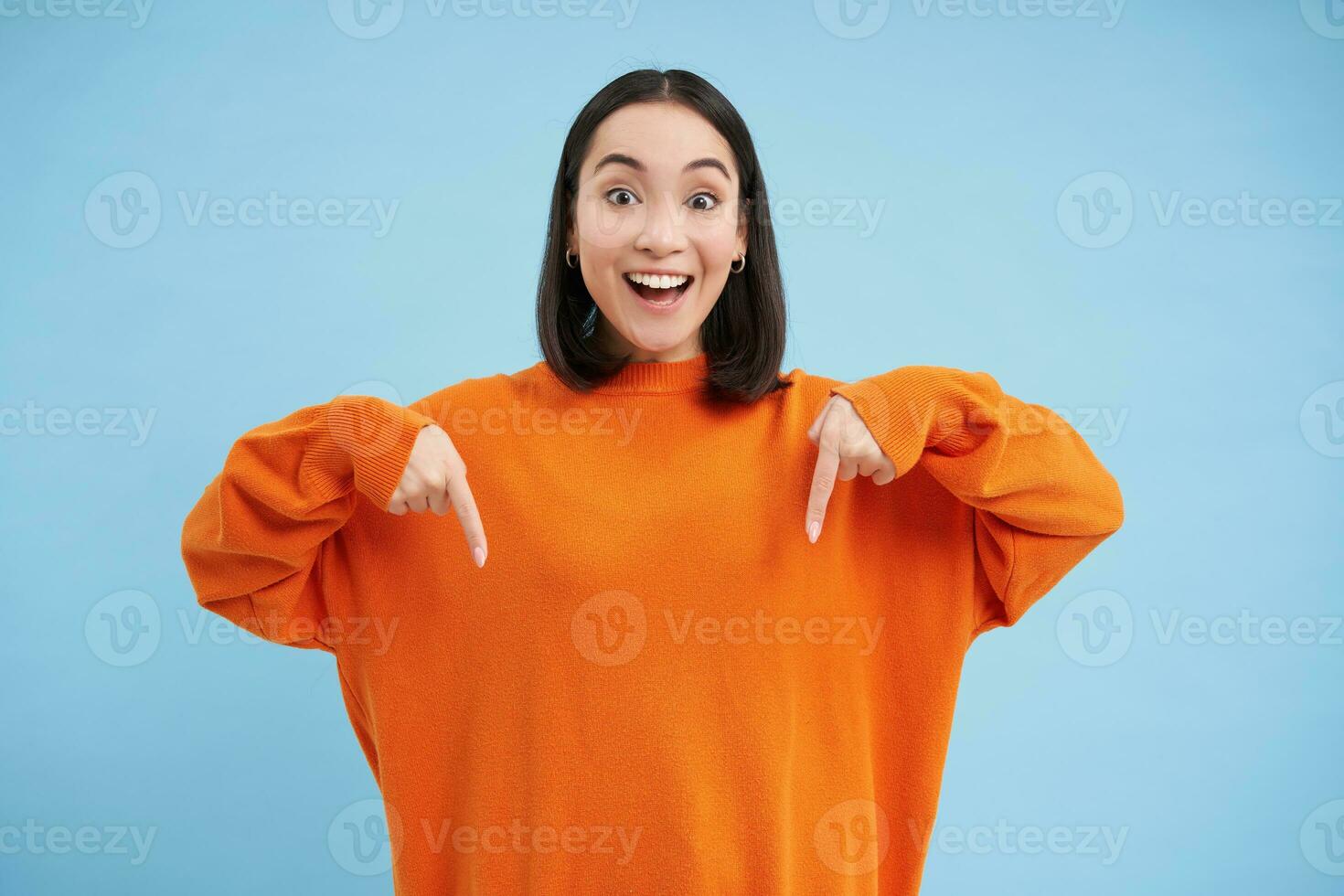 Cheerful asian woman shows advertisement below, points down with smiling face, inviting people to click on link, follow arrow, standing over blue background photo