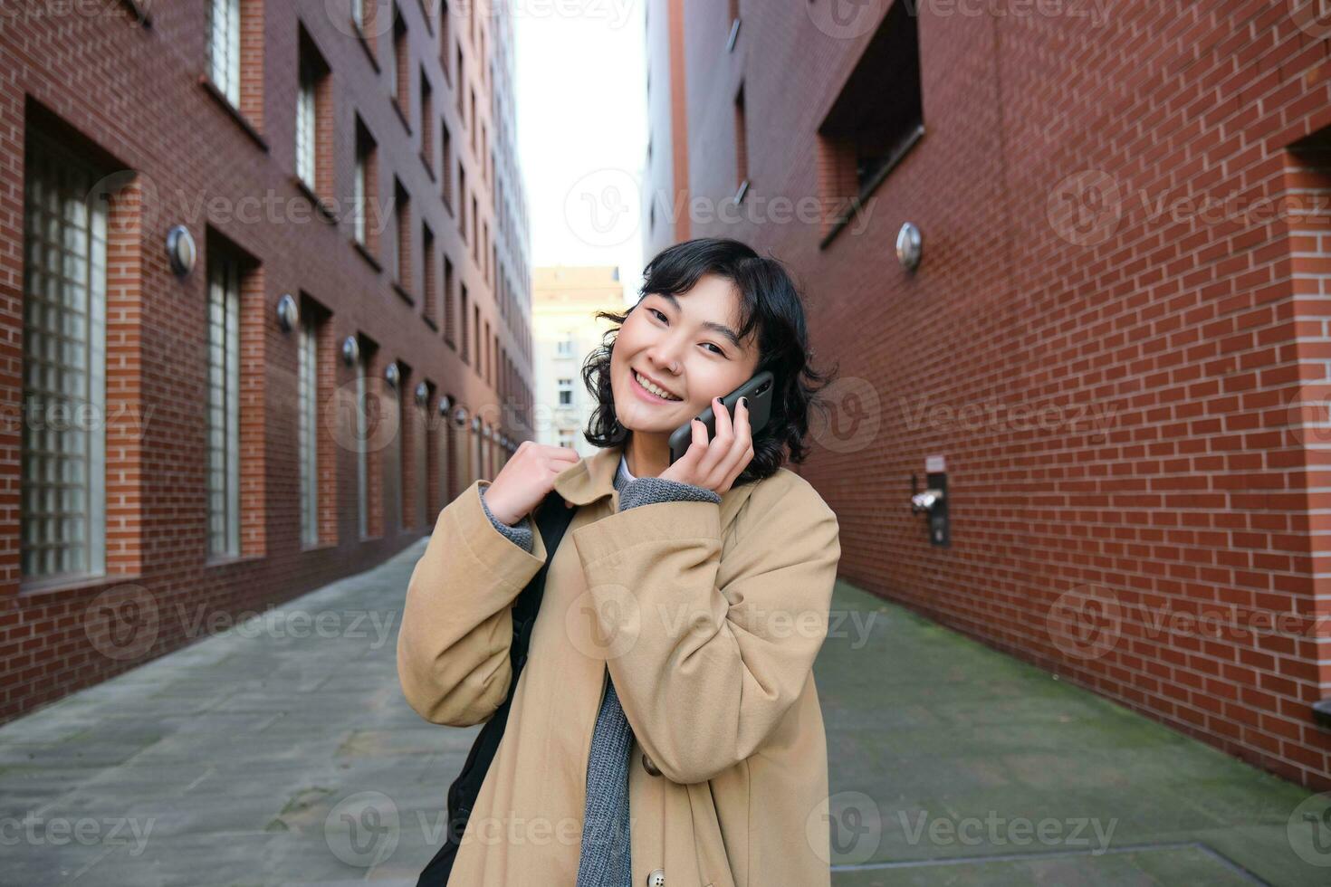 retrato de joven coreano mujer caminando abajo calle con teléfono, hablando con alguien, hace un llamar, tiene teléfono conversacion foto