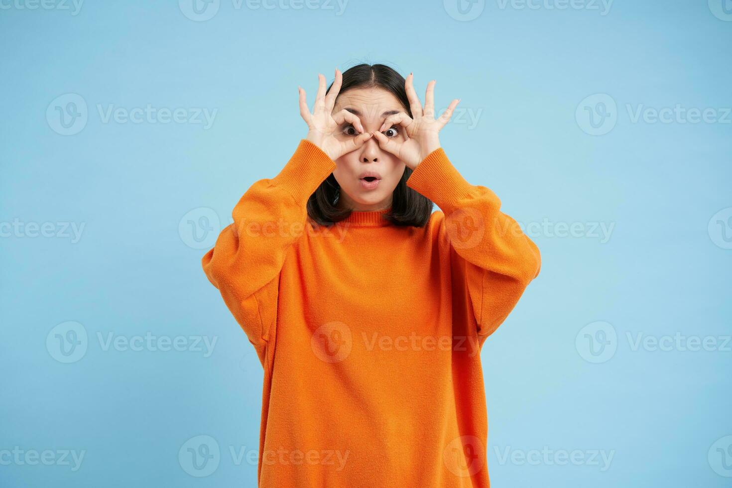 Wow, look at that. Portrait of amazed young asian woman, looks through hand glasses with impressed face expression, stands over blue background photo
