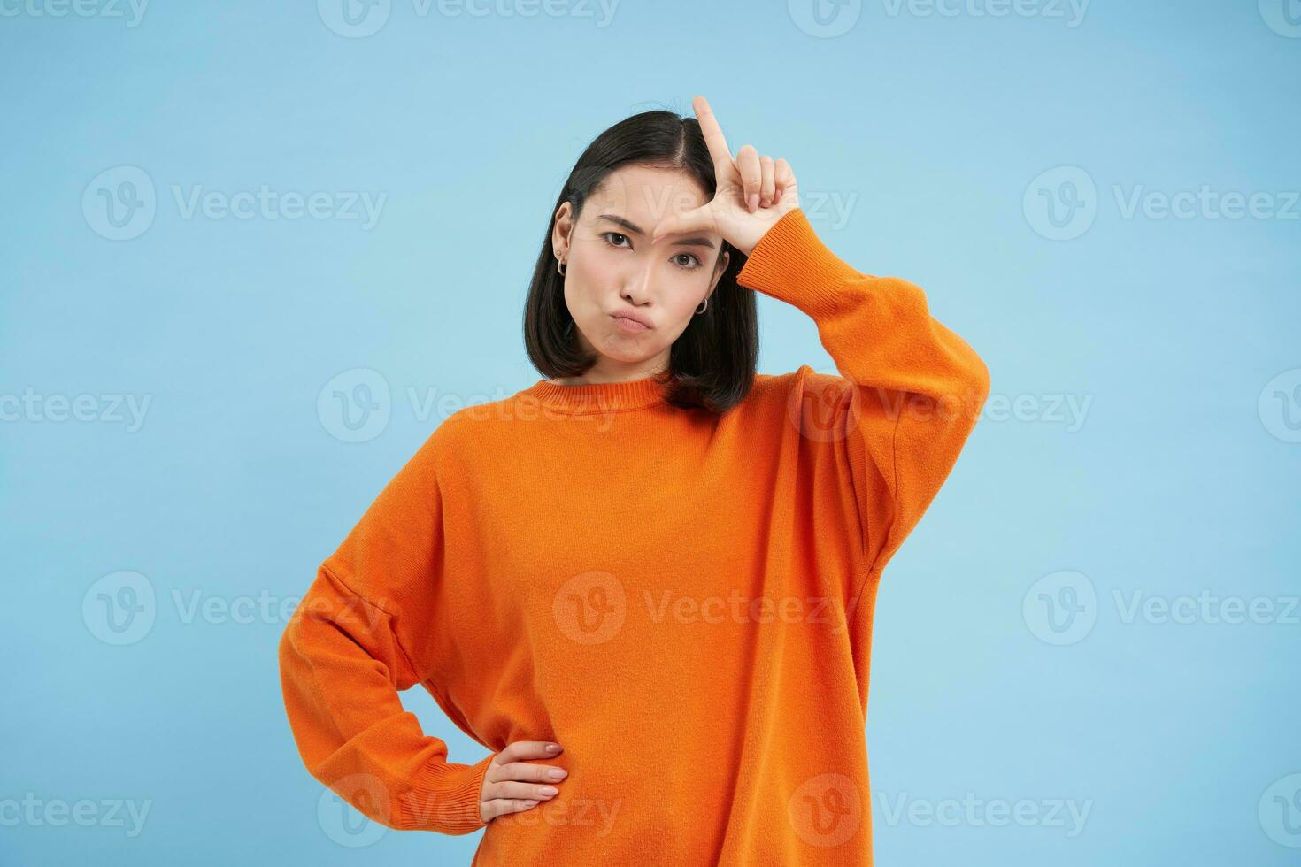 Losers. Young asian woman shows L letter on her forehead, stares sassy and confident at camera, stands over blue background photo