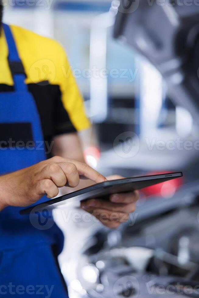 Precise serviceman in car service orders new engine for damaged vehicle using tablet. Employee looking for components on internet to replace old ones in malfunctioning automobile, close up photo