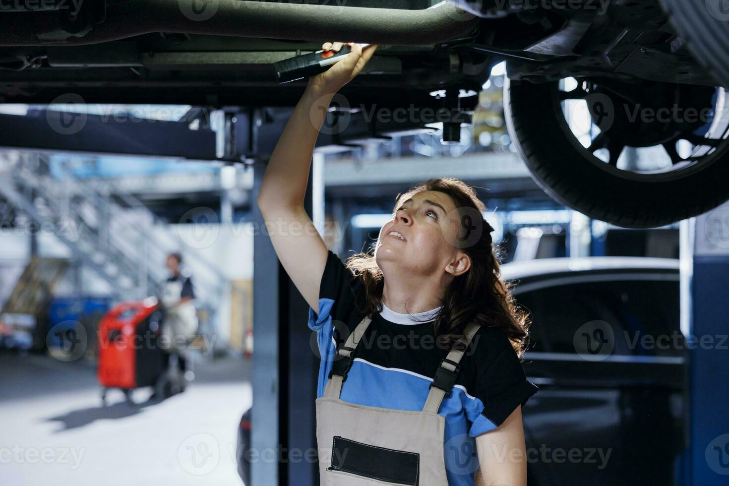 Meticulous mechanic working on suspended car in garage, checking motor during routine maintenance. Auto repair shop employee underneath vehicle, inspecting parts using work light photo