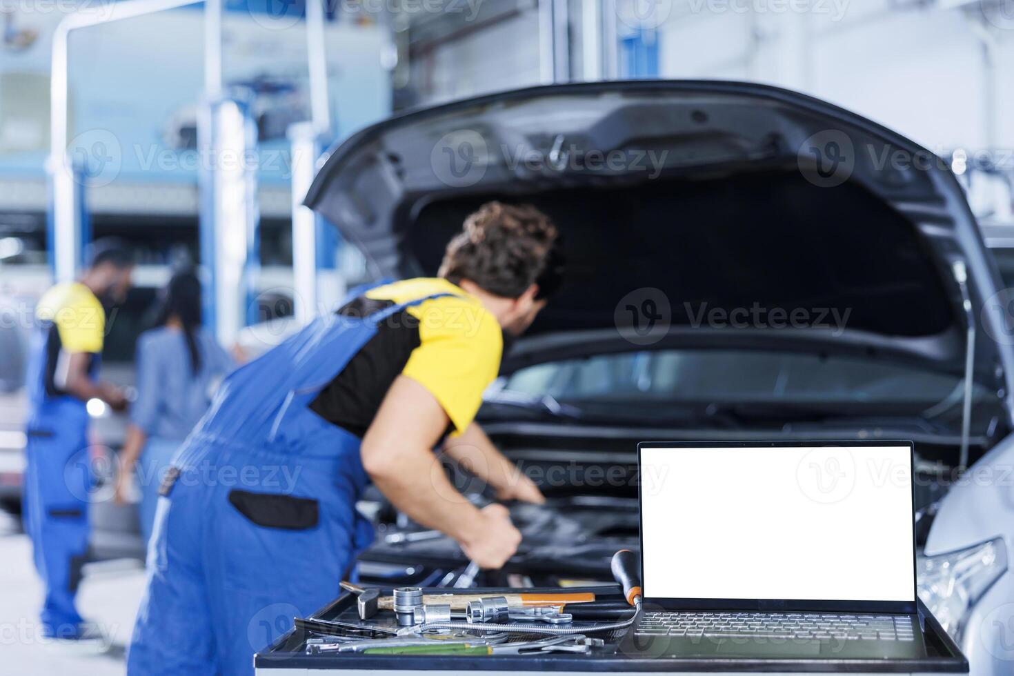 Mockup laptop on car service work station bench with technician in blurry background replacing engine. Isolated screen device next to qualified garage expert fixing client automobile photo