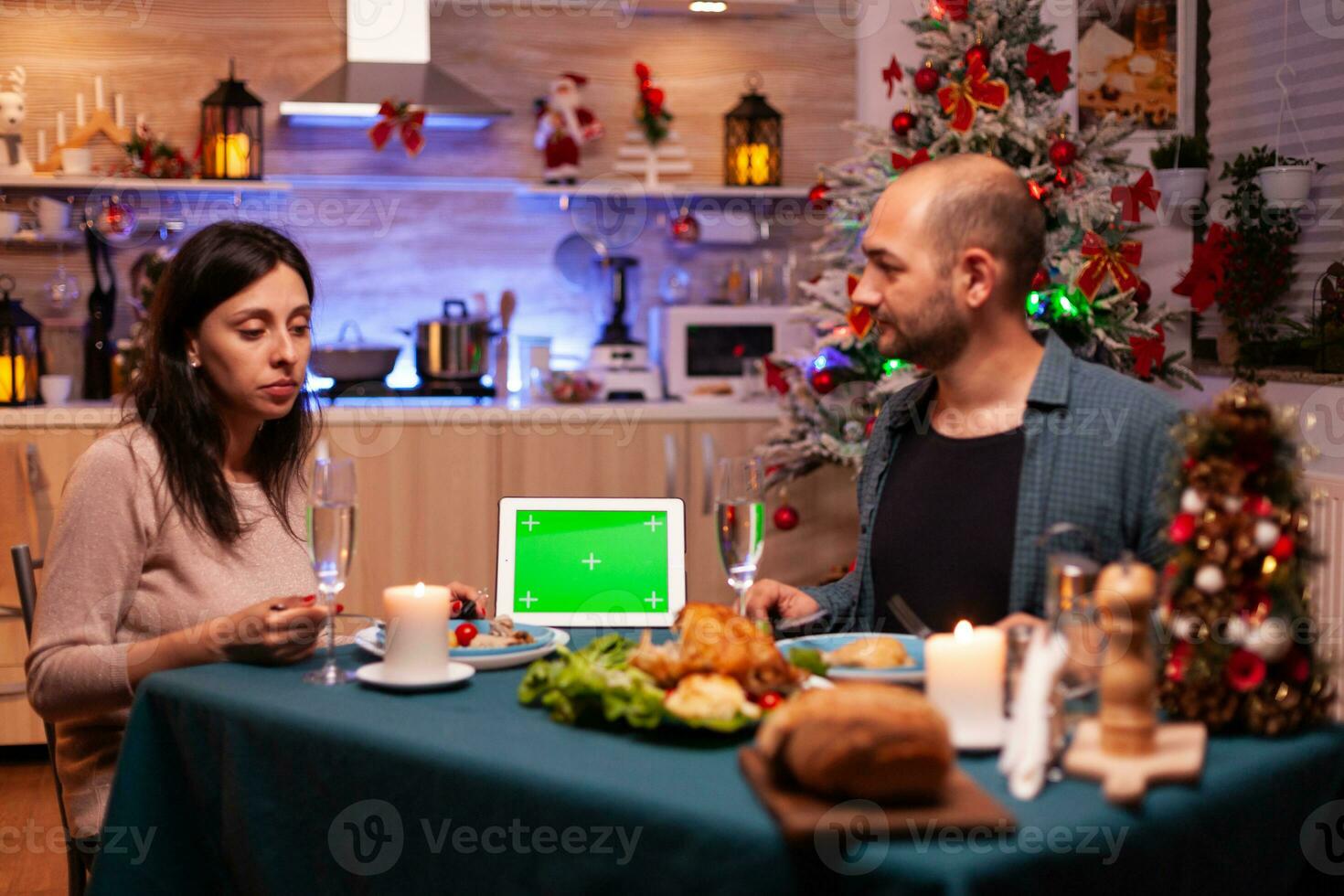 Cheerful family enjoying xmas dinner in x-mas decorated kitchen celebrating christmas holiday. Green screen mock up chroma key tablet computer with isolated display standing on table photo