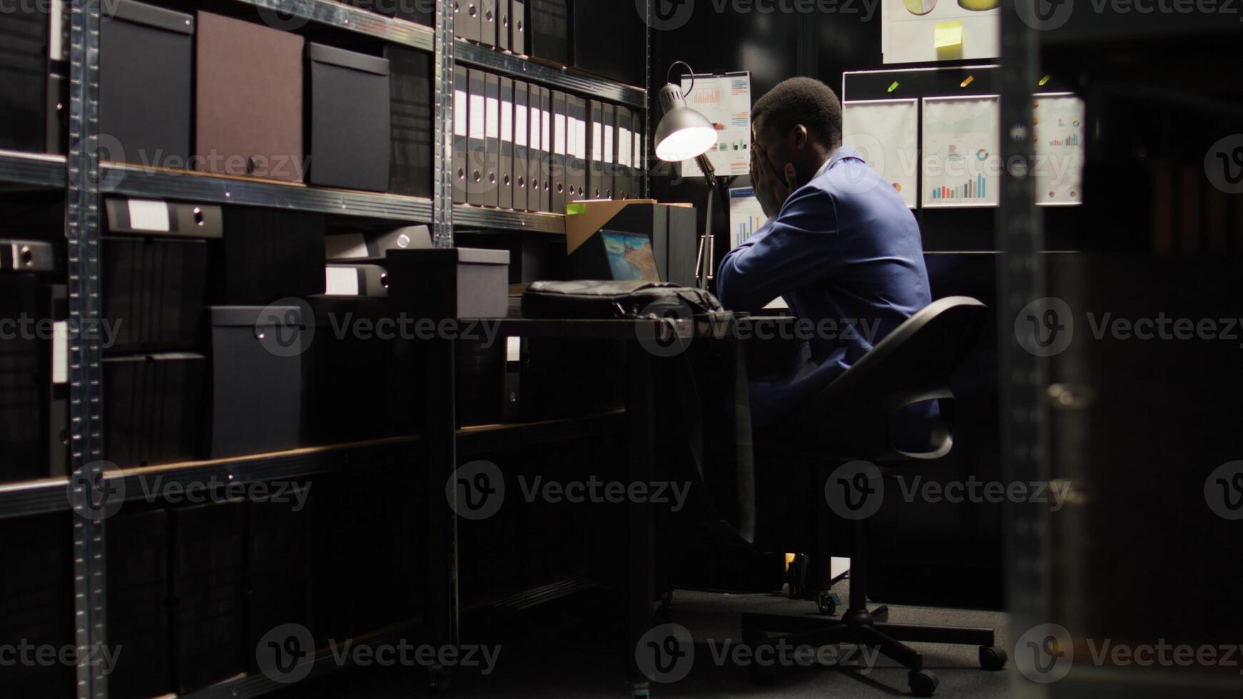Surveillance experienced african american law enforcement official walks into office with a laptop bag. Detective sets laptop down on desk and starts looking through suspect files and documents. photo