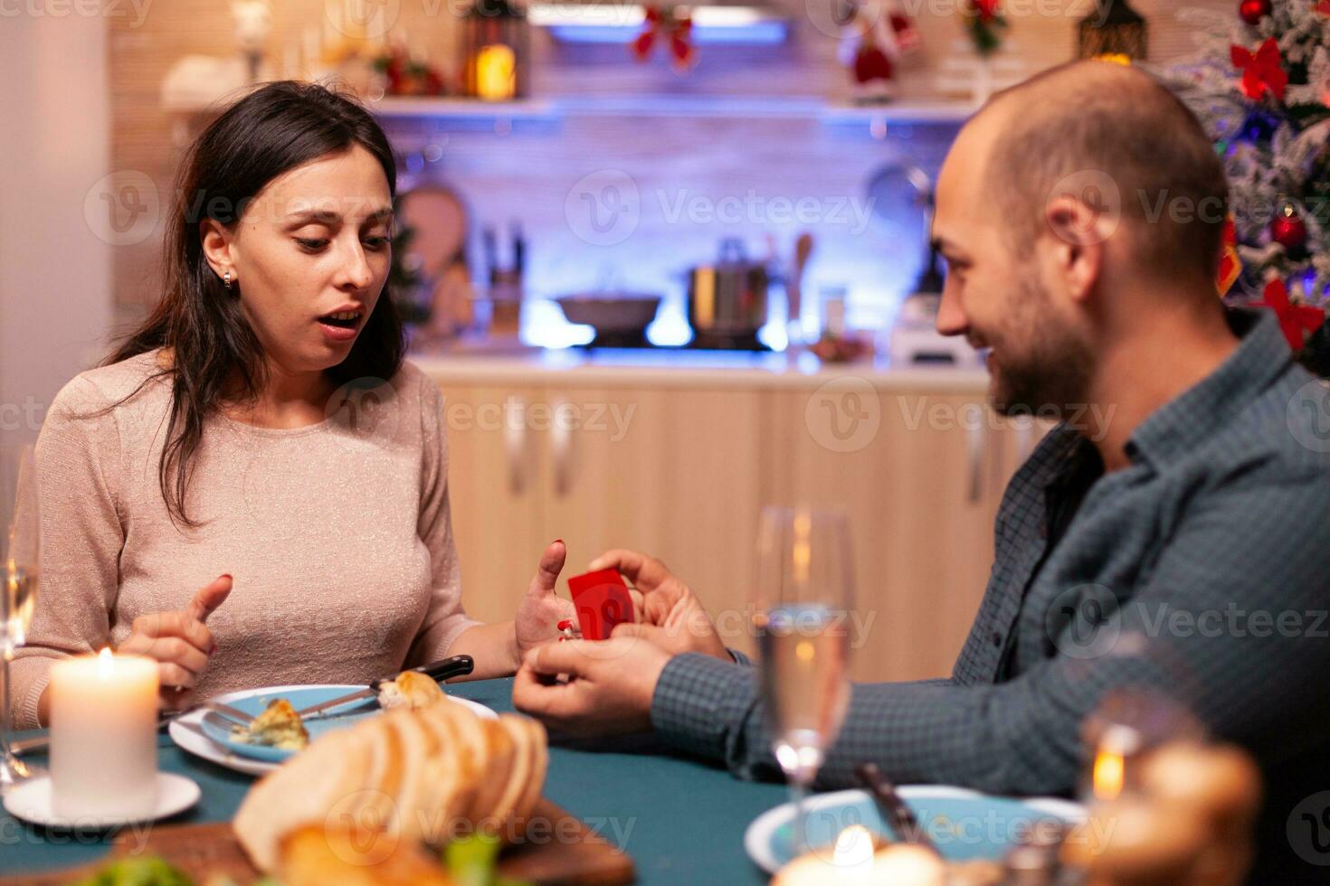 Happy man surpsing girlfriend with diamond luxury engagement ring enjoying christmas holiday together sitting at dinning table in xmas decorated kitchen. Married couple enjoying winter season photo