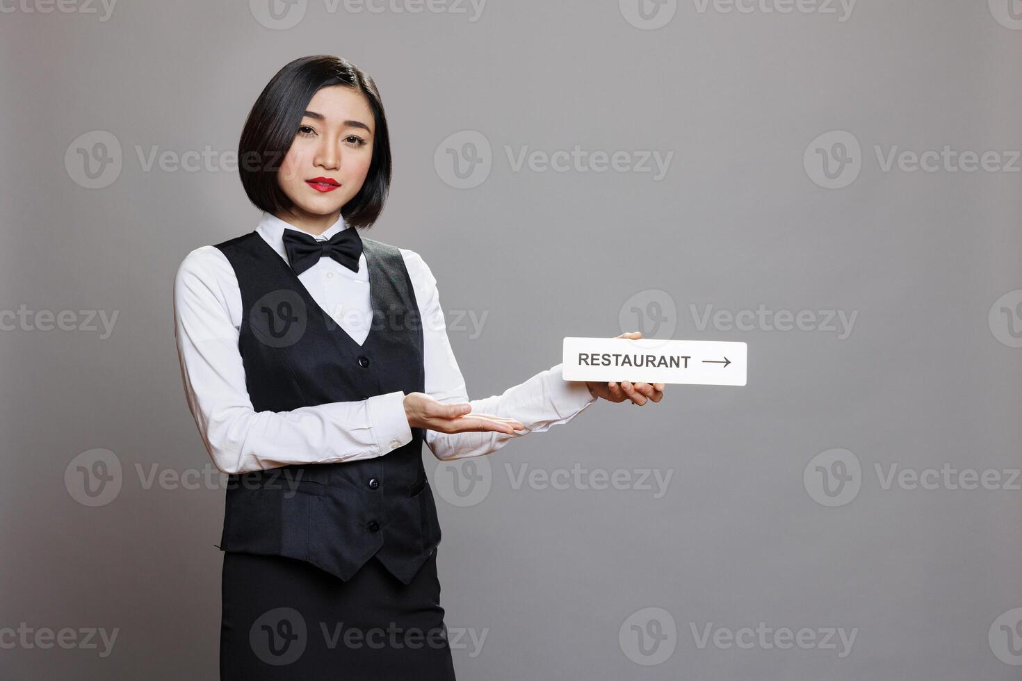 Asian waitress holding restaurant direction signage and looking at camera while posing in studio. Hospitality service woman employee in uniform showing cafeteria pointer steel sign portrait photo