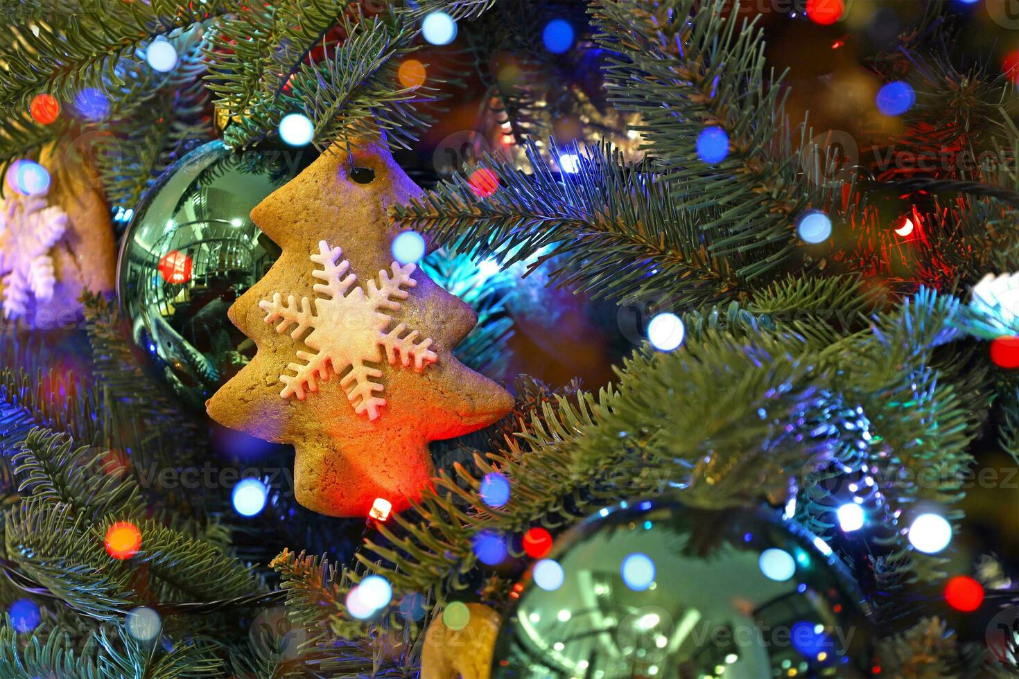 Christmas tree decorated with gingerbread cookies, green balls, light garland. Gingerbread cookie in form of Xmas tree with white snowflake. Holiday colorful blurred bokeh lights background. photo