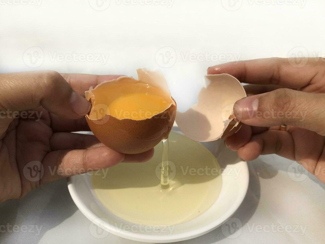 A woman's hand cracks an egg to separate the egg white and yolk and the egg shell in the background. photo