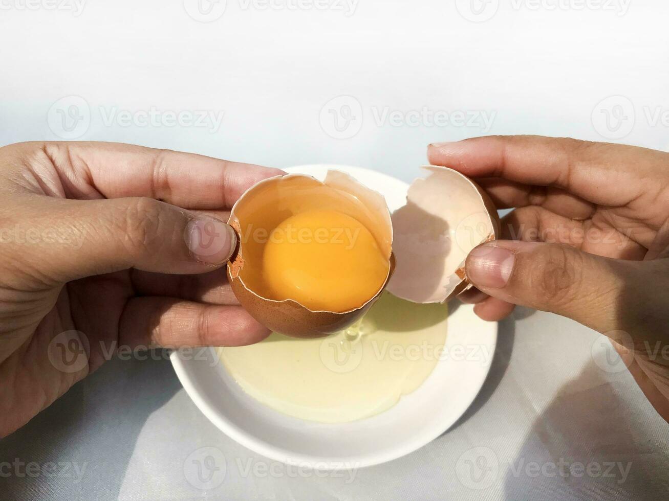 A woman's hand cracks an egg to separate the egg white and yolk and the egg shell in the background. photo