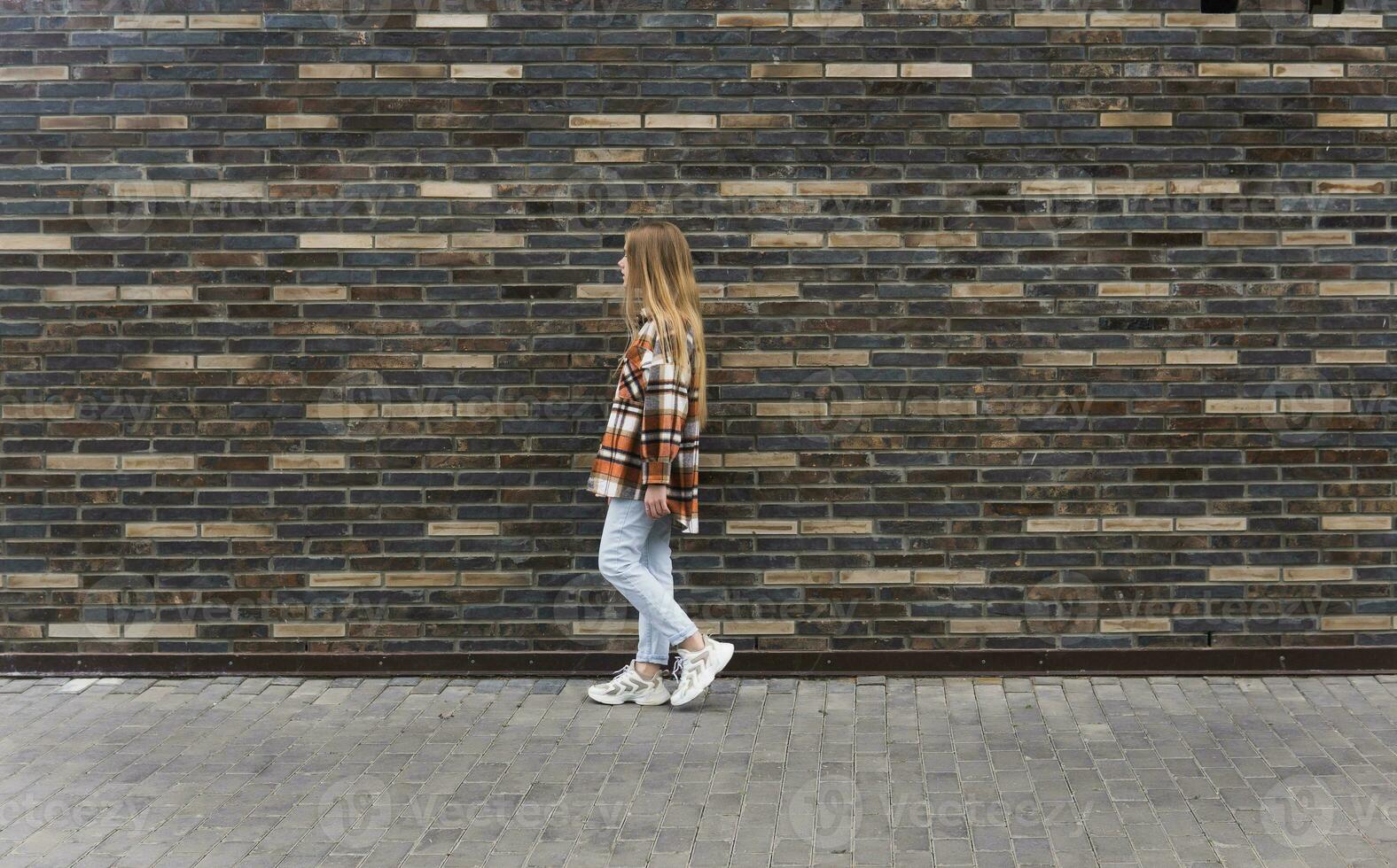 young woman walks down the street against a brick wall photo