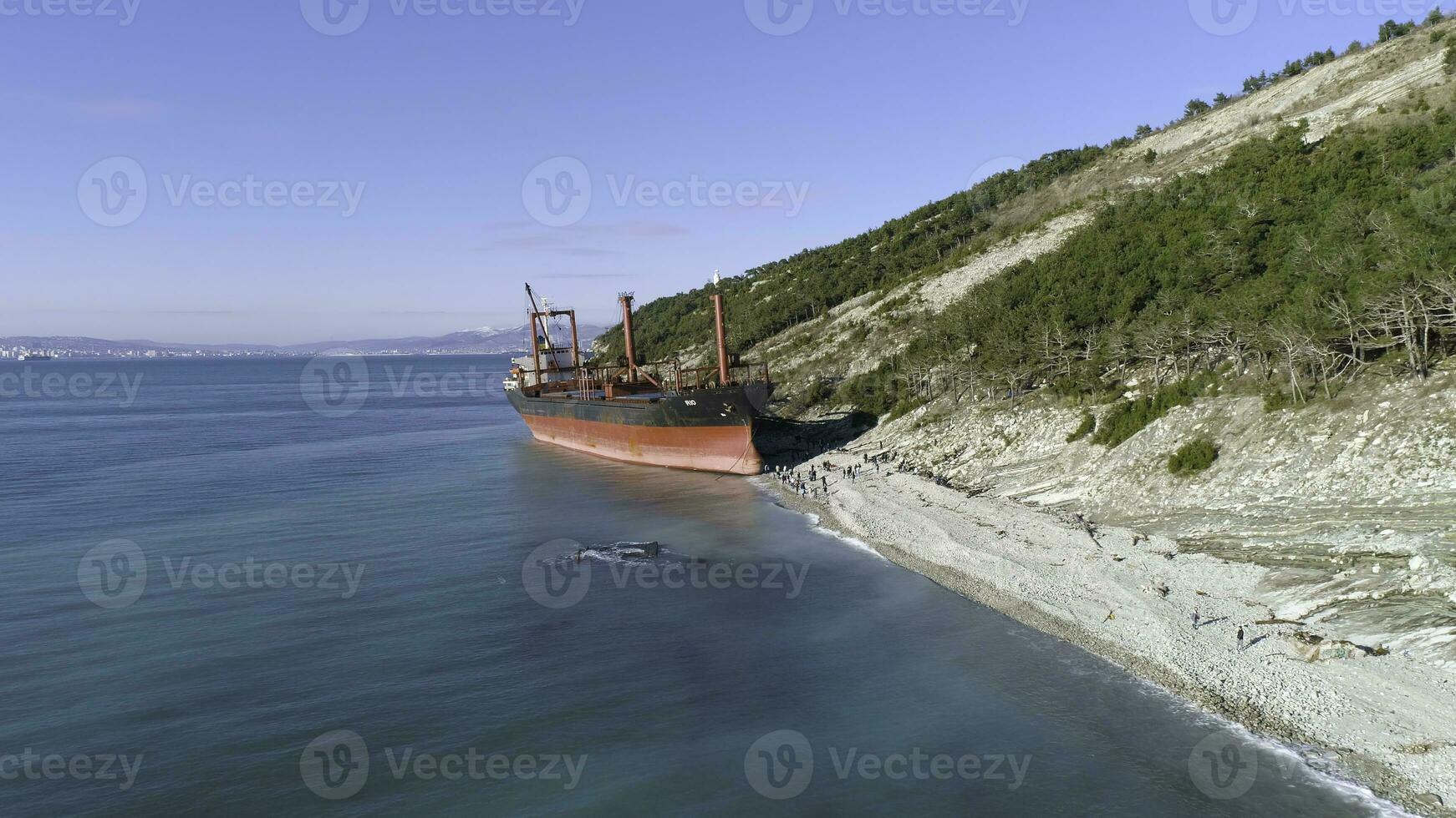 Aerial for an empty industrial ship moored near sea shore with many people walking on a beach. Maritime cargo vessel standing near green trees slope in a summer sunny day. photo
