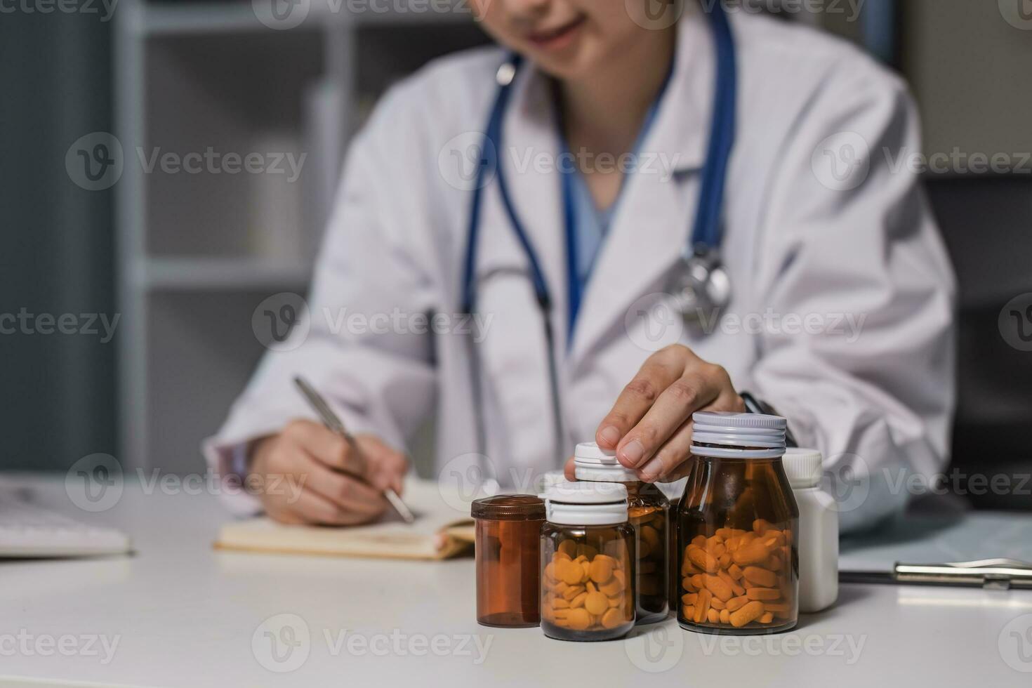 Asian female medic doctor gp therapist wear white coat holding pills bottle in hand writing medical prescription sitting at work desk prescribing pharmacy medicine concept. Closeup view photo