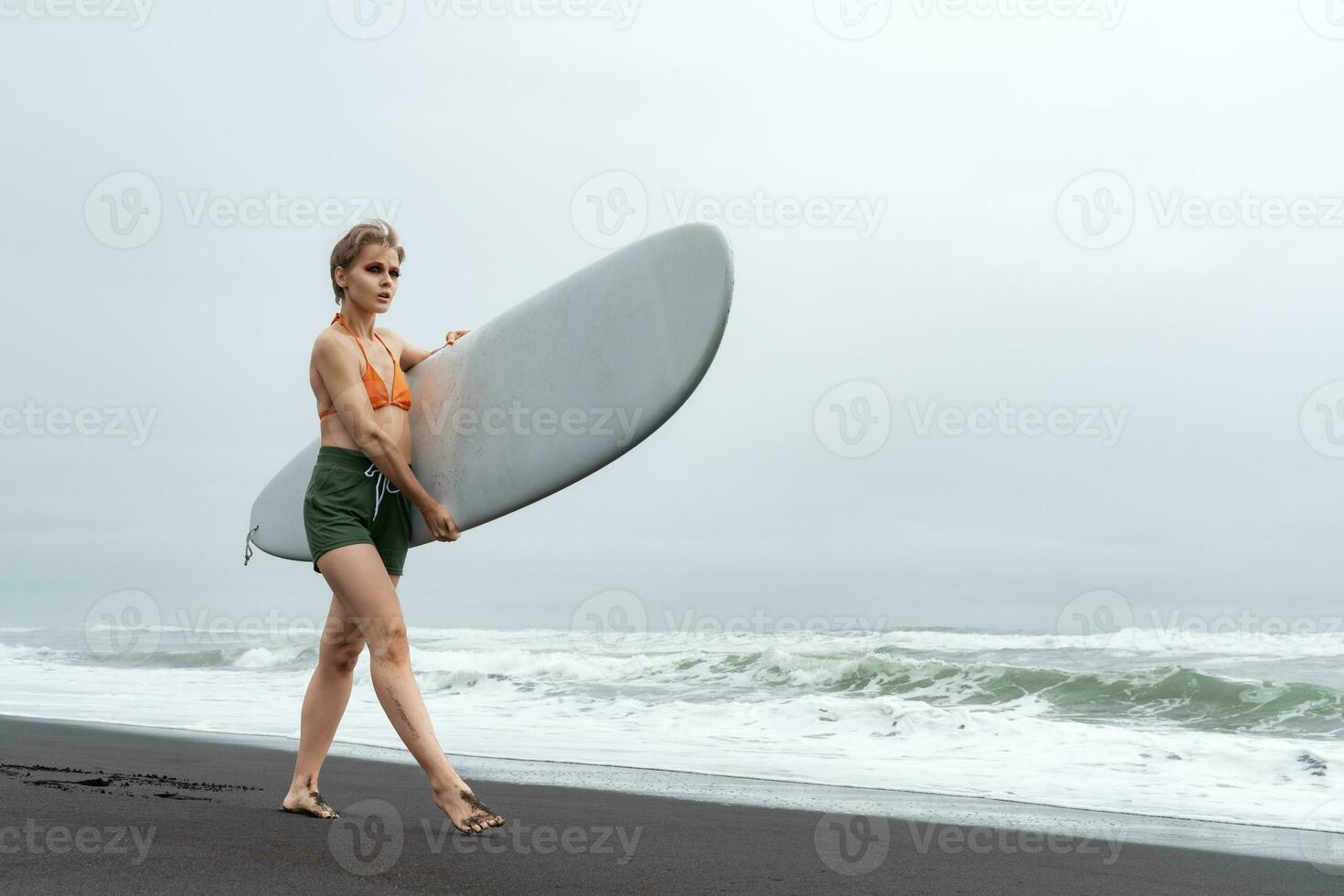 Female surfer walks along black sand beach while carrying surfboard against backdrop of ocean waves photo