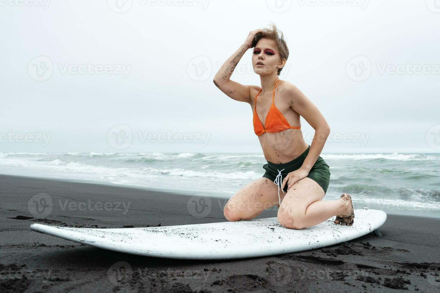 Woman surfer posing on knees on surfboard, looking at camera on beach on background of ocean waves photo