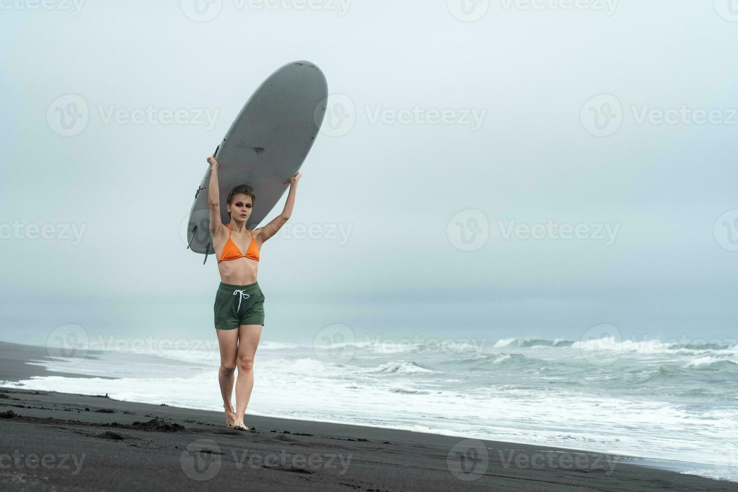 Woman surfer walking on beach and carrying white surfboard on head against background of sea waves photo