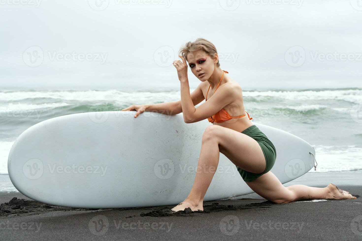 Female surfer stands on knee, stretches one leg forward on beach and holds surfboard with one hand photo