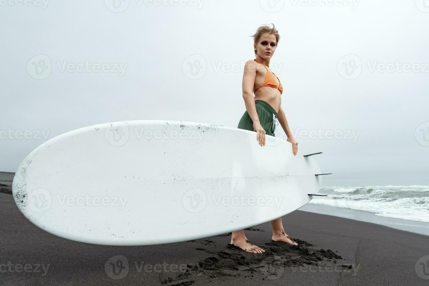 Female surfer standing on black sandy beach holding white surfboard against background of sea waves photo
