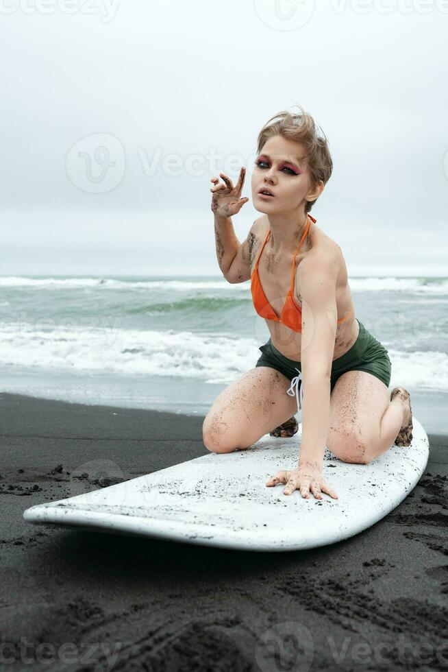 Sensuality woman surfer strikes pose on knees atop surfboard. Sports fashion model looking at camera photo